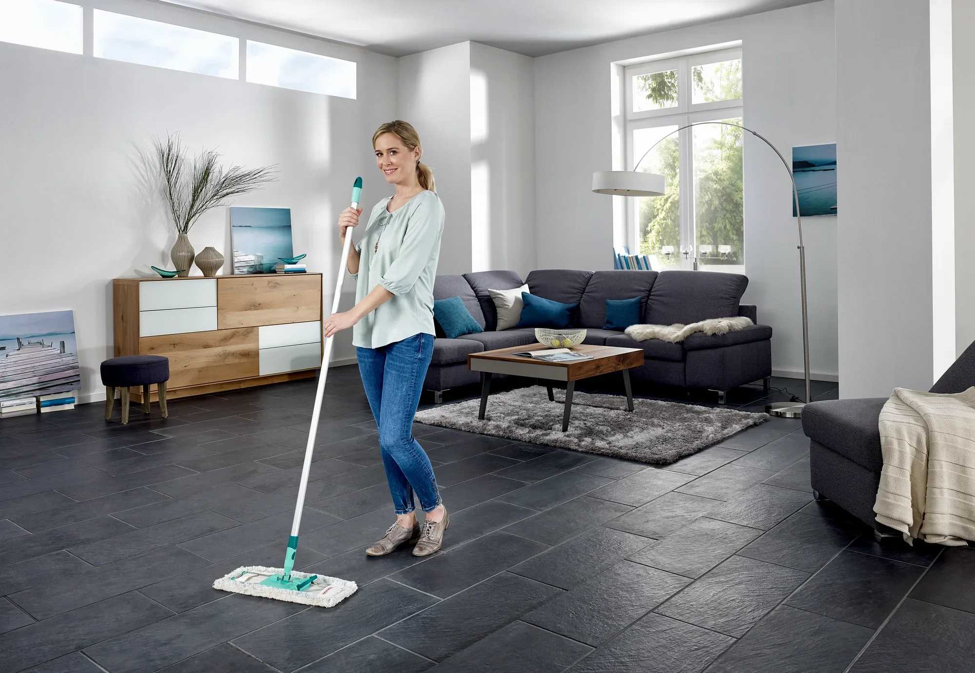 Una donna pulisce il pavimento di un moderno soggiorno con il lavapavimenti Profi XL. La stanza è luminosa e arredata con stile, con grandi finestre e mobili eleganti.