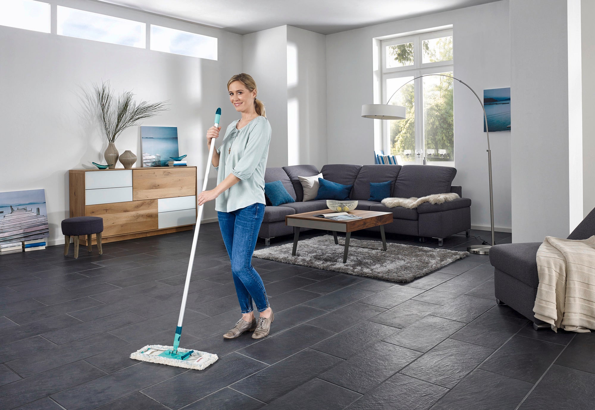Eine Frau wischt den Boden in einem modernen Wohnzimmer mit dem Bodenwischer Profi XL. Der Raum ist hell und stilvoll eingerichtet, mit großen Fenstern und eleganten Möbeln.