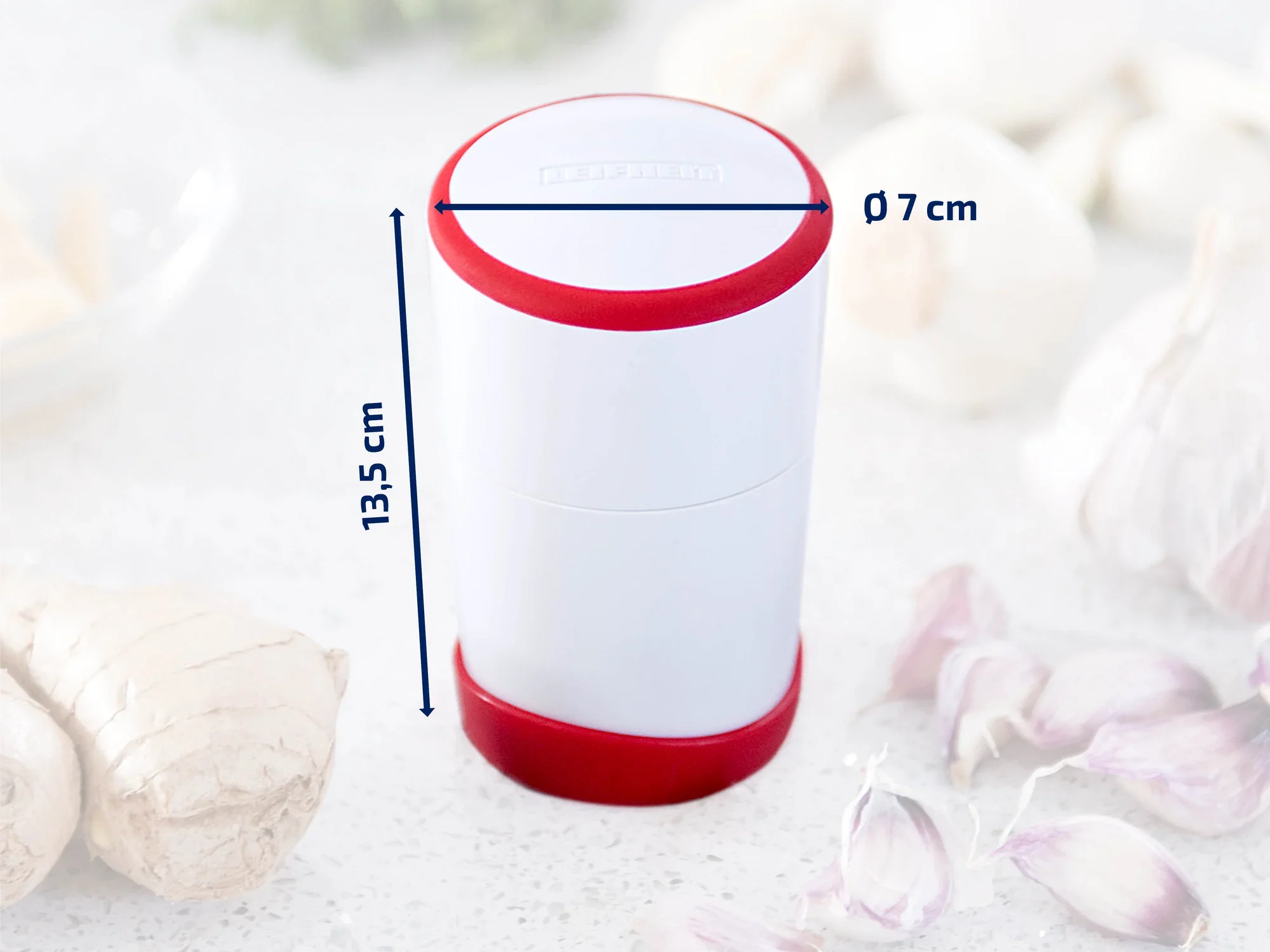 Een witte, cilindervormige knoflooksnijder met rode accenten, 13,5 cm hoog en 7 cm in diameter, staat op een keukenblad. Omringd door knoflooktenen en gember.