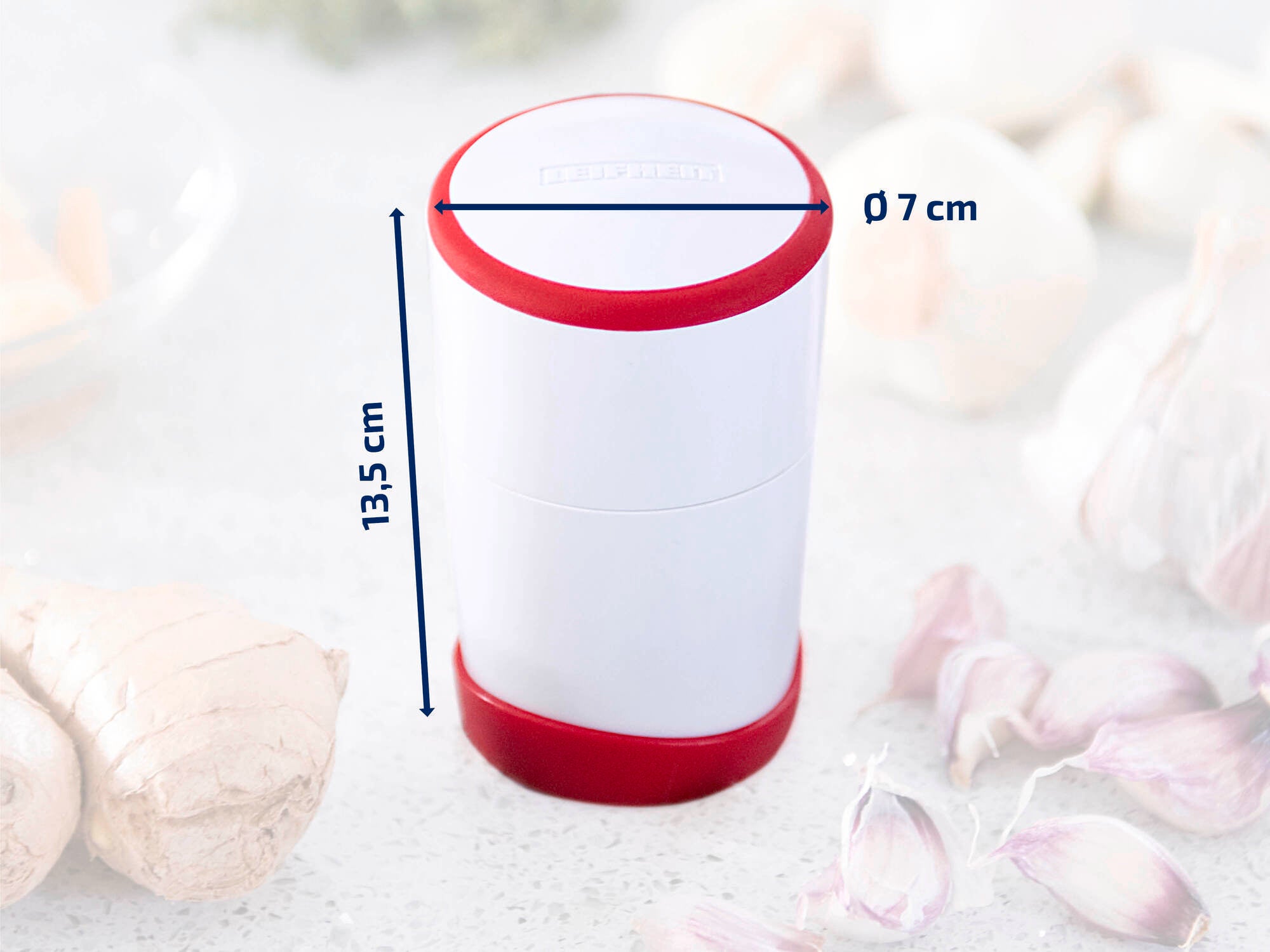 Un coupe-ail cylindrique blanc avec des accents rouges, de 13,5 cm de haut et 7 cm de diamètre, est posé sur un plan de travail de cuisine. Entouré de gousses d'ail et de gingembre.