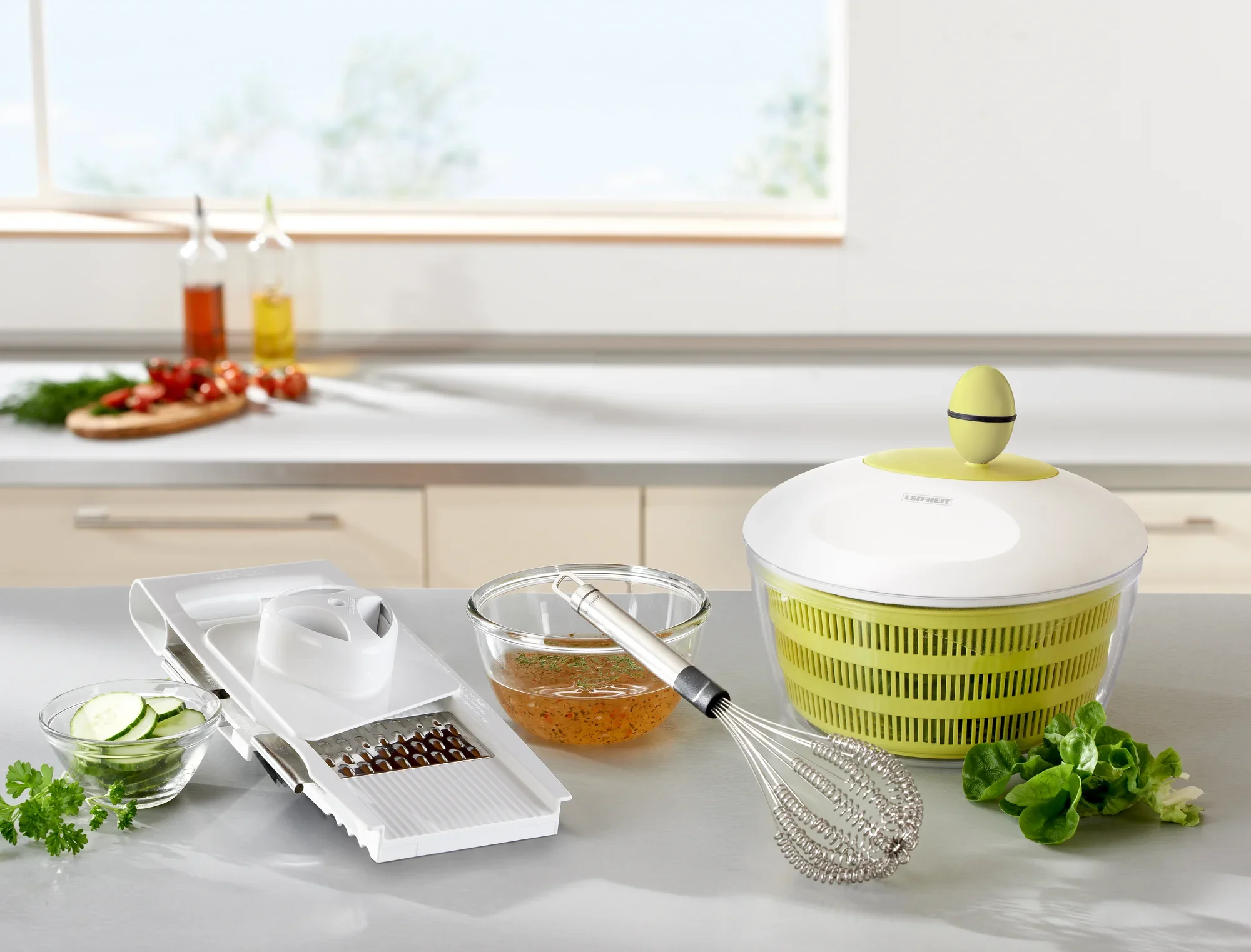 Essoreuse à salade Trend dans une cuisine moderne. À côté de l'essoreuse, on aperçoit un fouet, une râpe avec des rondelles de concombre et des herbes fraîches. Idéale pour les salades fraîches.
