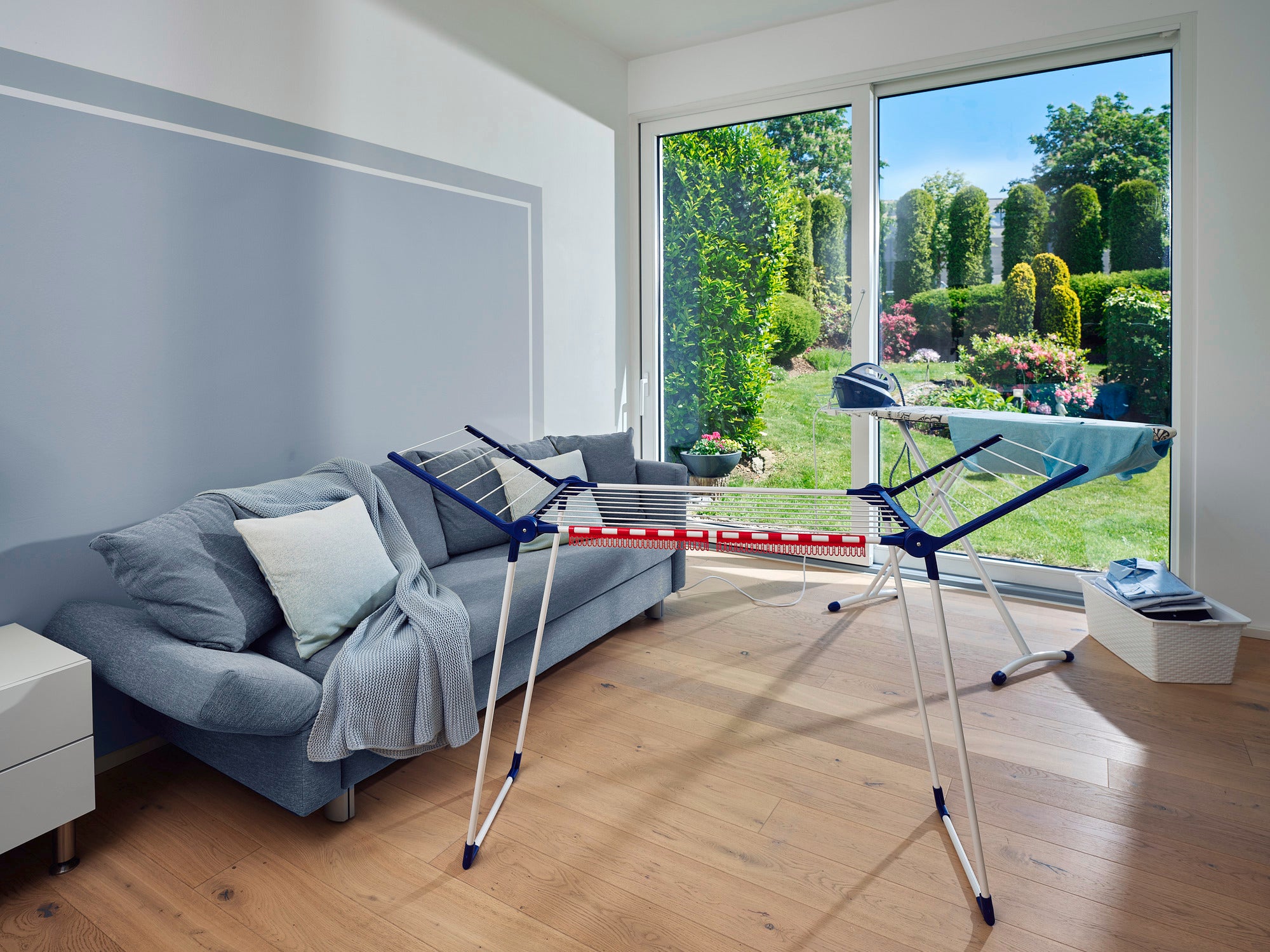 Ein Standtrockner Pegasus 150 Solid Slim steht in einem modernen Wohnzimmer mit Blick auf einen gepflegten Garten. Der Trockner ist kompakt und ideal für kleine Räume.