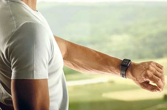 Ein Mann in einem weissen T-Shirt traegt eine schwarze Smartwatch, sein Arm ausgestreckt vor einer gruenen, verschwommenen Landschaft.