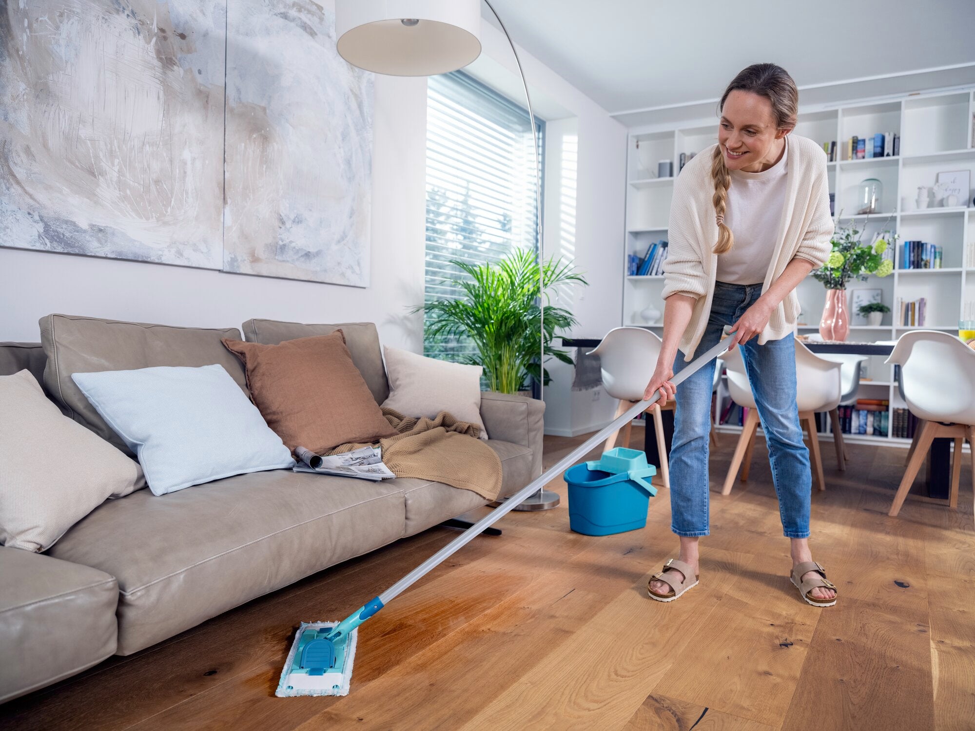 Eine Frau wischt den Holzboden eines modernen Wohnzimmers mit dem Bodenwischer POWER CLEAN. Im Hintergrund sind ein Sofa, Pflanzen und ein Bücherregal zu sehen. Ideal für gründliche Reinigung.