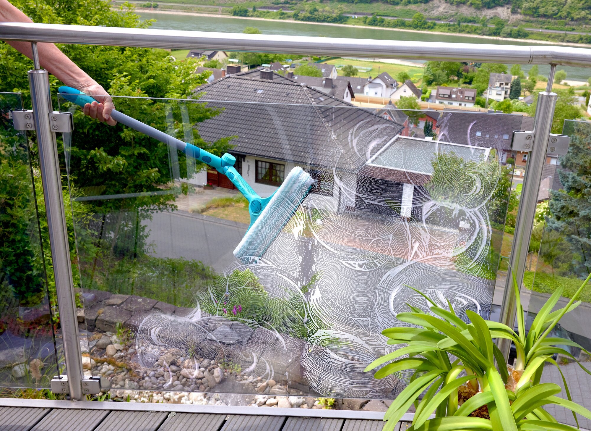 Ein Mensch reinigt mit dem Leifheit 4in1 FlexReach Fensterwischer ein Glasgeländer auf einem Balkon. Im Hintergrund sind Häuser und eine grüne Landschaft zu sehen.