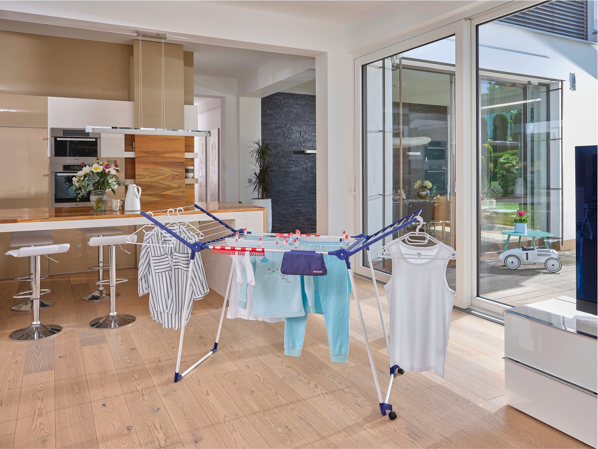 Ein Standtrockner Pegasus 200 Solid Deluxe Mobile steht in einem modernen Wohnzimmer. Er ist mit verschiedenen Kleidungsstücken behängt und bietet viel Platz zum Trocknen.