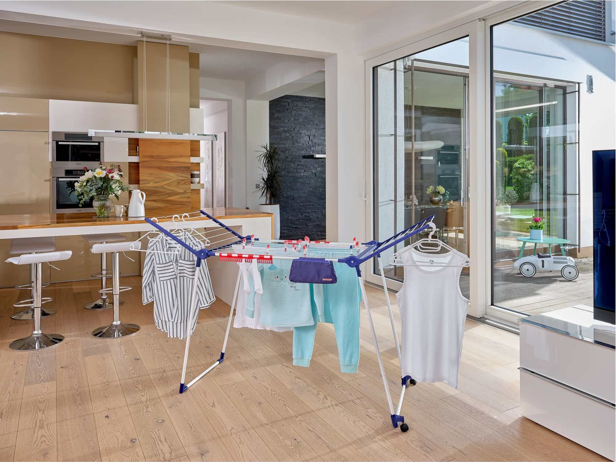 Suszarka Pegasus 200 Solid Deluxe Mobile stoi w nowoczesnym salonie. Jest zawieszona na różnych ubraniach i oferuje dużo miejsca do suszenia.