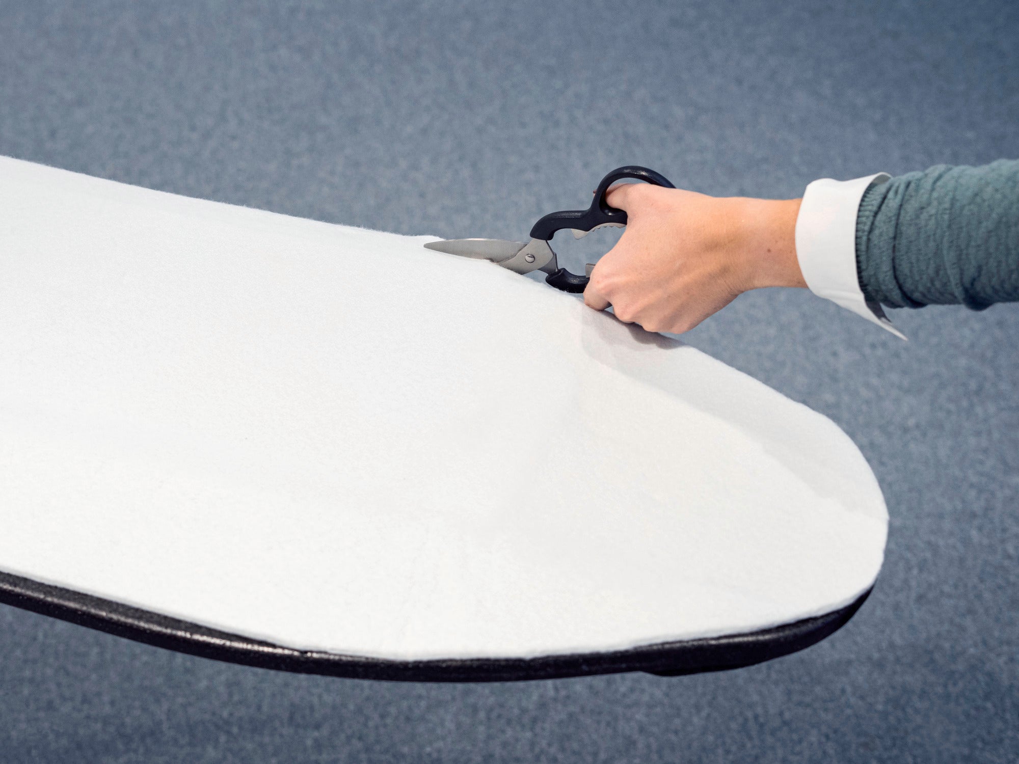 Eine Person schneidet mit einer Schere die Bügeltischpolsterung passend zu. Die weiße Polsterung wird präzise zugeschnitten, um optimal auf den Bügeltisch zu passen.