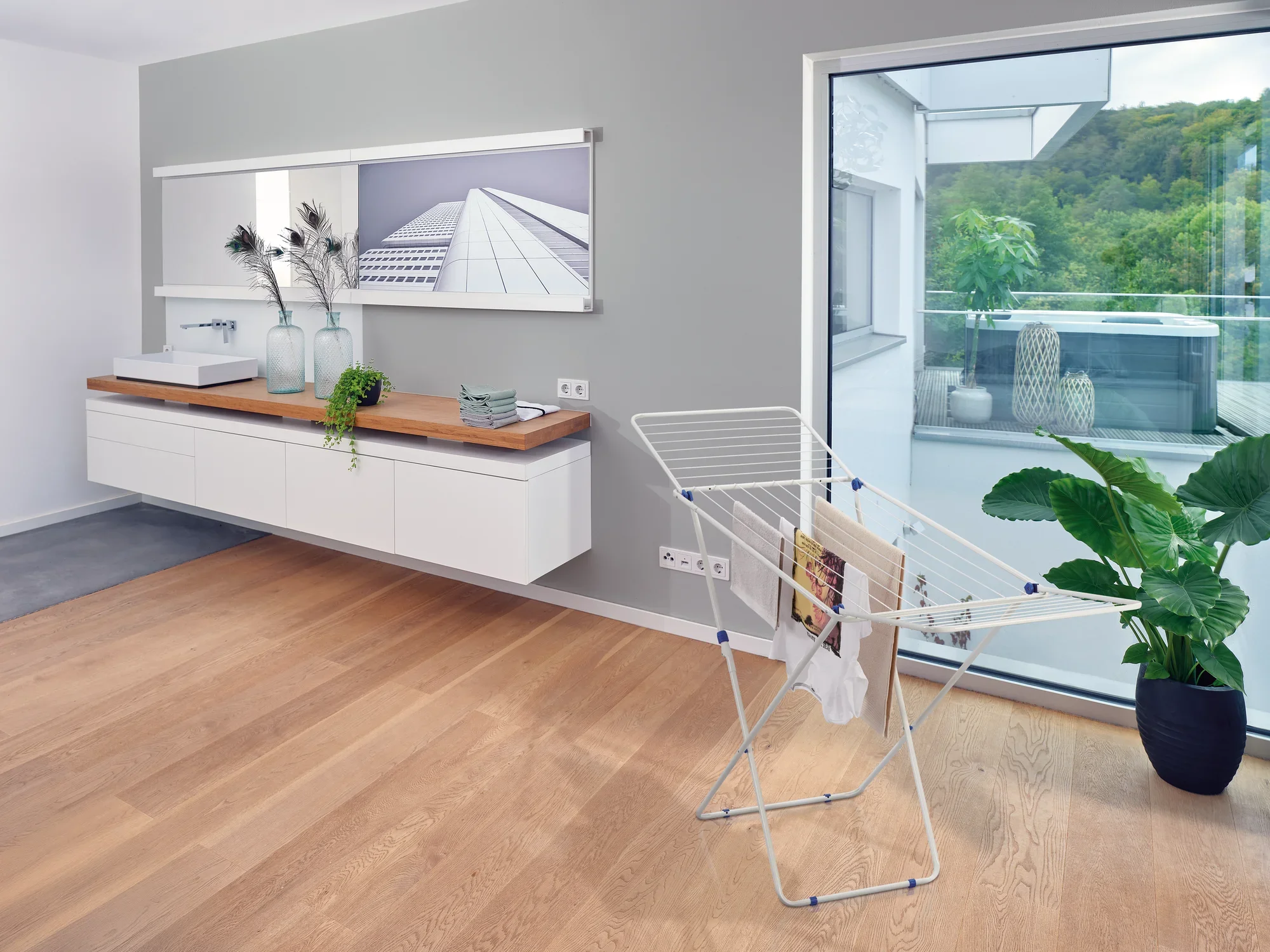 Wolnostojąca suszarka bębnowa Classic 200 Easy stoi w nowoczesnym, jasnym pomieszczeniu z drewnianą podłogą. W tle widać umywalkę i duże okno z widokiem na zieleń.