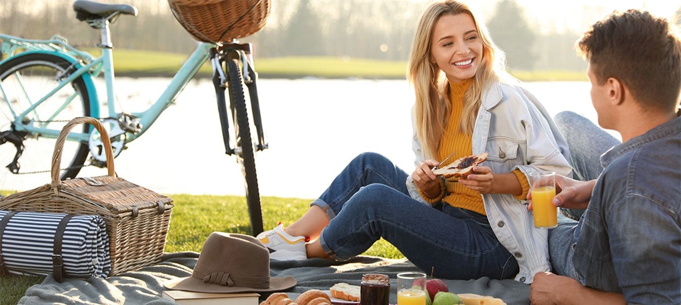 Picknick im Grünen Ein Paar sitzt auf einer Decke beim Picknick am See, umgeben von Essen, einem Fahrrad und einer idyllischen Landschaft.