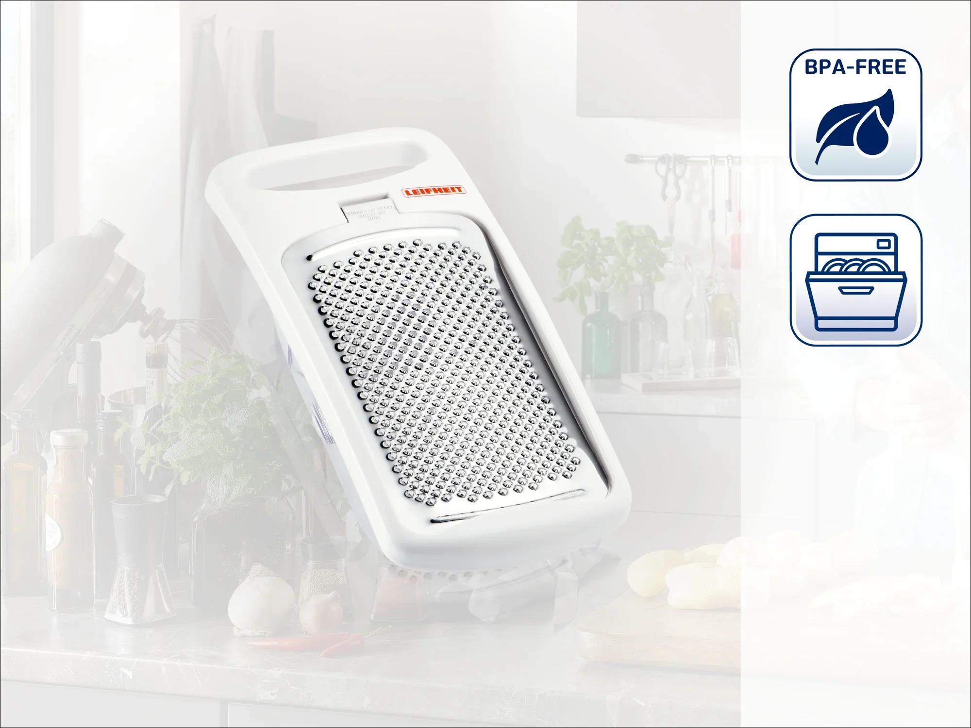 Une râpe blanche Leifheit est posée sur un plan de travail dans une cuisine moderne. Des ustensiles de cuisine sont visibles à l'arrière-plan. Le produit est sans BPA et passe au lave-vaisselle.