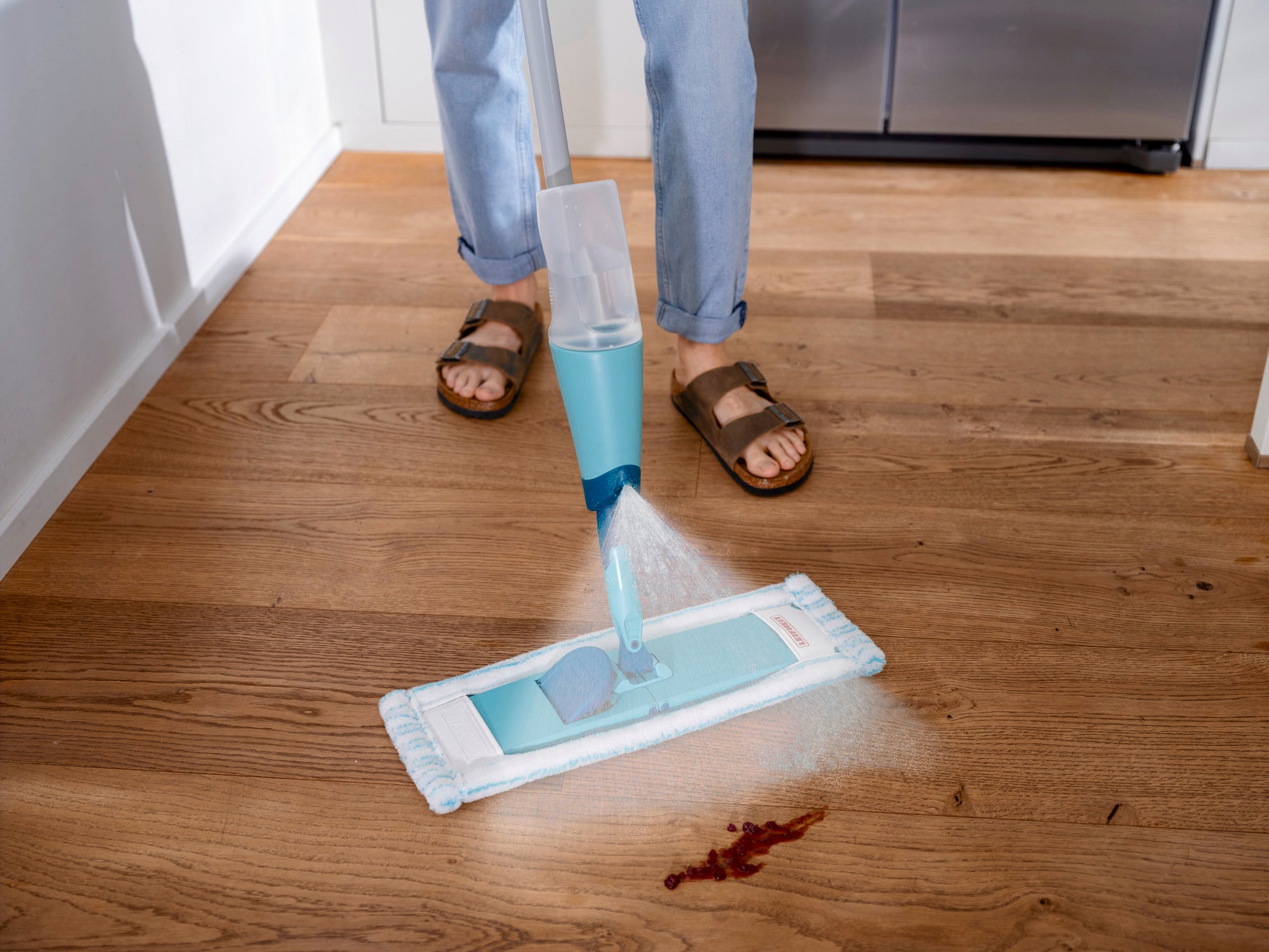 Un balai-serpillière Easy Spray XL est utilisé sur un parquet pour nettoyer une petite tache. Le balai-serpillière pulvérise de l'eau devant lui pendant qu'une personne le manie.