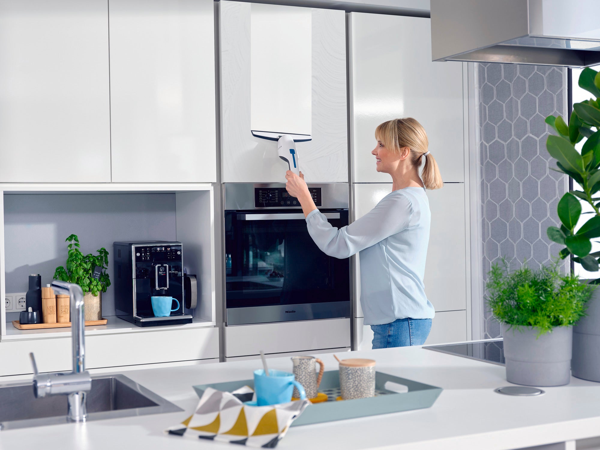 A woman cleans a window in a modern kitchen with the Nemo window vacuum cleaner. The vacuum cleaner is compact and efficient, ideal for everyday use in the home.