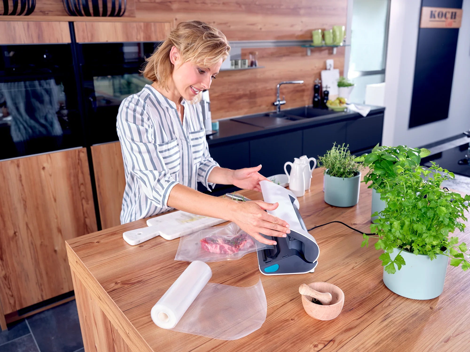 Une femme utilise l'appareil de mise sous vide Vacu Power 300 dans une cuisine moderne. Des sachets sous vide et des herbes fraîches dans un pot sont posés sur la table. Idéal pour la conservation des aliments.