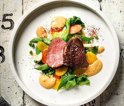 Ein Teller mit zwei Rinderfilets, umgeben von Erbsen, Karotten, Pak Choi und einer cremigen Sosse auf weissem Hintergrund.
