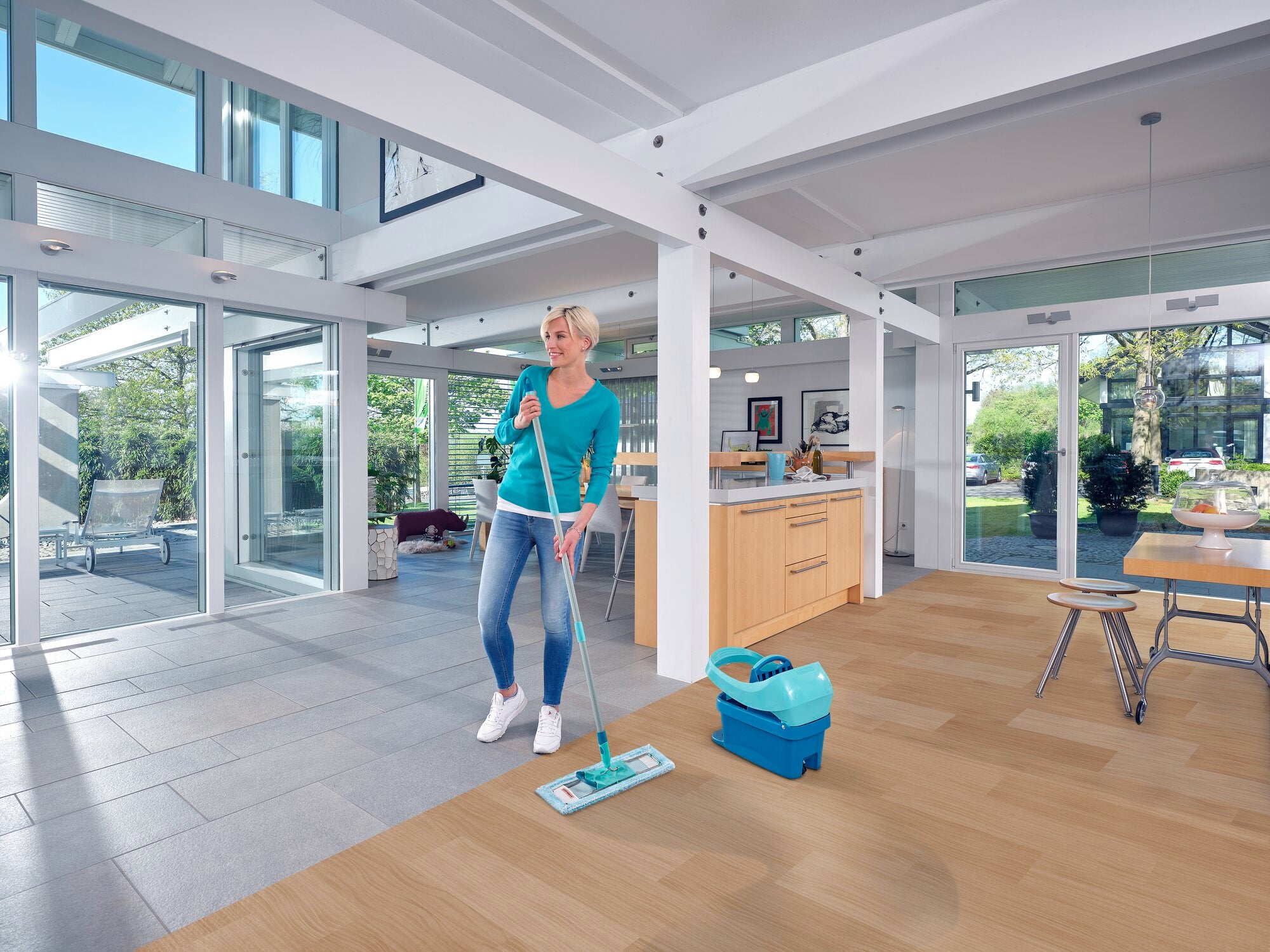 Eine Frau wischt den Boden eines modernen, lichtdurchfluteten Wohnzimmers mit dem Bodenwischer Profi XL. Der Raum hat große Fenster und einen Holzboden, der mit dem Wischer gereinigt wird.