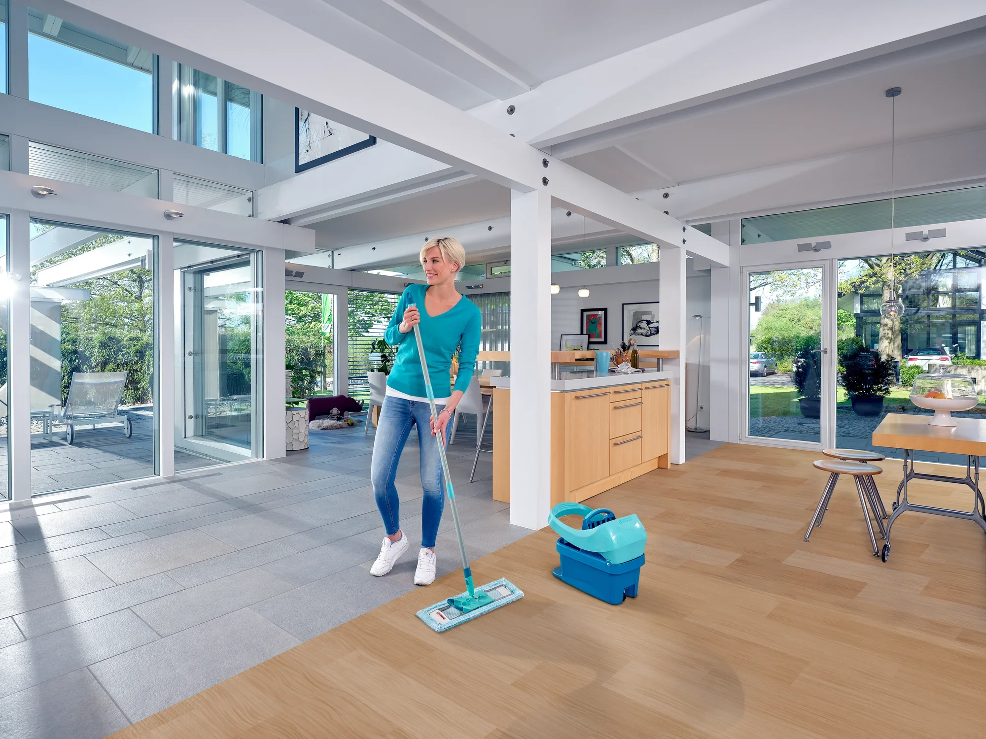 Una donna pulisce il pavimento di un soggiorno moderno e luminoso con il lavapavimenti Profi XL. La stanza ha grandi finestre e un pavimento in legno che viene pulito con il lavapavimenti.