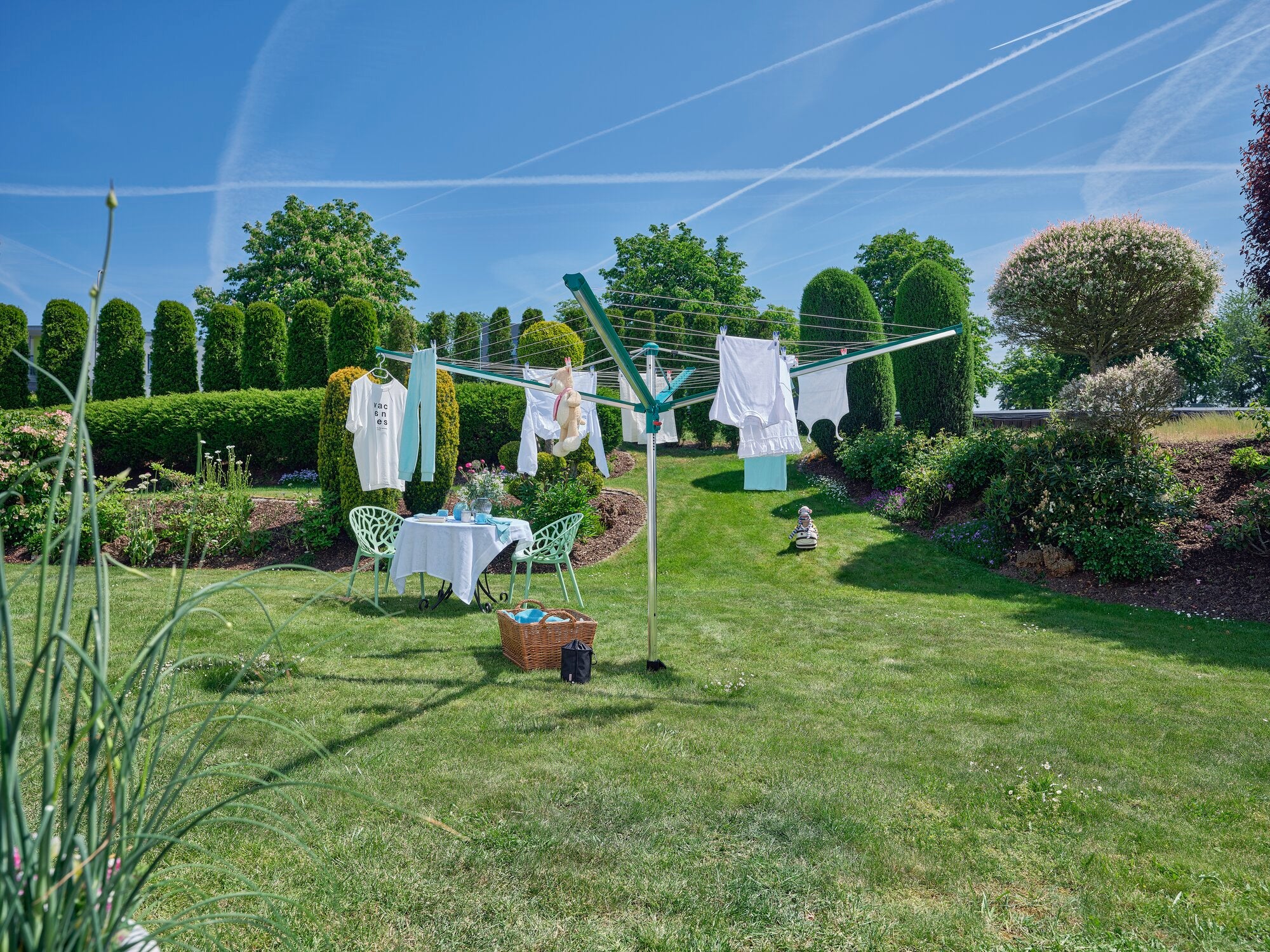 Wäschespinne Linomatic 500 Deluxe in einem sonnigen Garten. Die Wäschespinne ist mit frisch gewaschener Kleidung behängt. Im Hintergrund sind grüne Hecken und Bäume zu sehen.