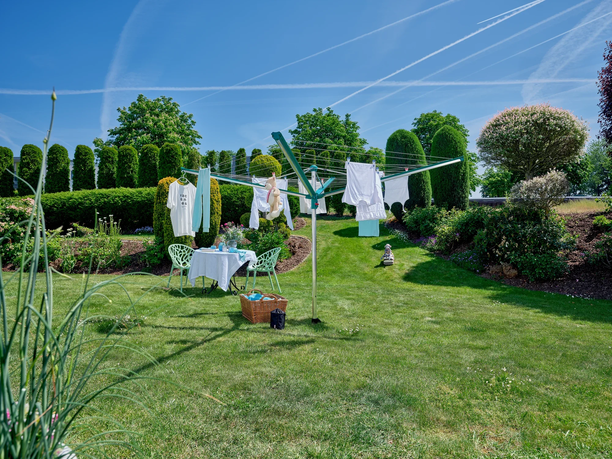 Suszarka obrotowa Linomatic 500 Deluxe w słonecznym ogrodzie. Suszarka obrotowa jest zawieszona na świeżo wypranych ubraniach. W tle widać zielone żywopłoty i drzewa.