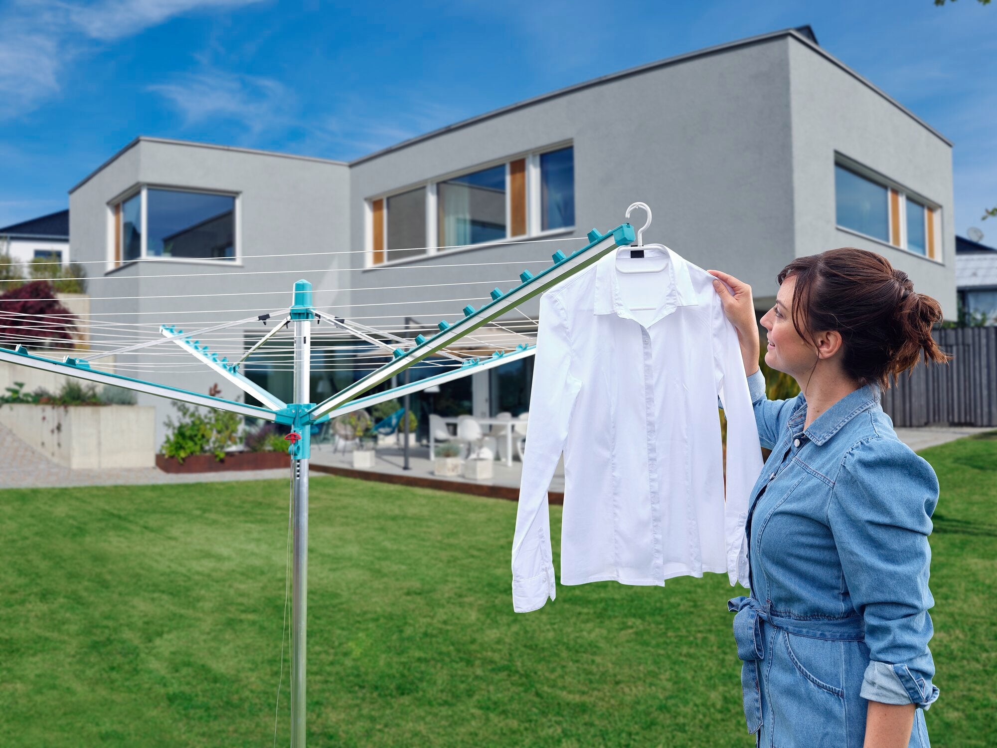 Een vrouw hangt een wit overhemd aan het Leifheit Linomatic 500 droogrek in een groene tuin. Op de achtergrond is een modern huis met grote ramen te zien.