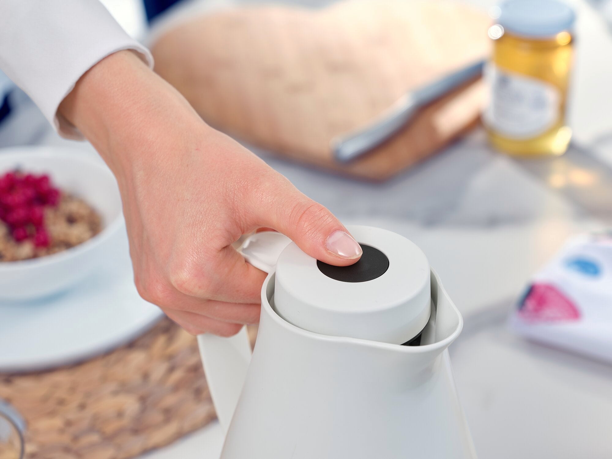 Harmonic vacuum jug in use. A hand presses the lid of the white jug to open it. Breakfast items such as muesli and honey can be seen in the background.