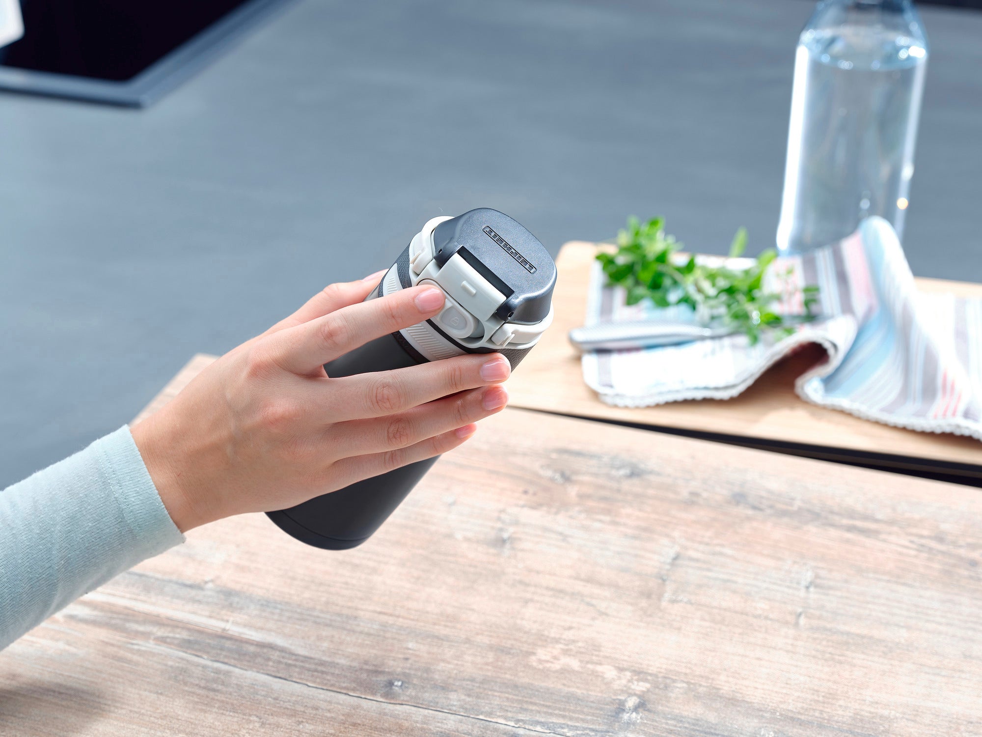 Une tasse isotherme Flip d'une contenance de 350 ml en noir est tenue dans une main. À l'arrière-plan, on aperçoit un verre d'eau, un torchon et des herbes fraîches.
