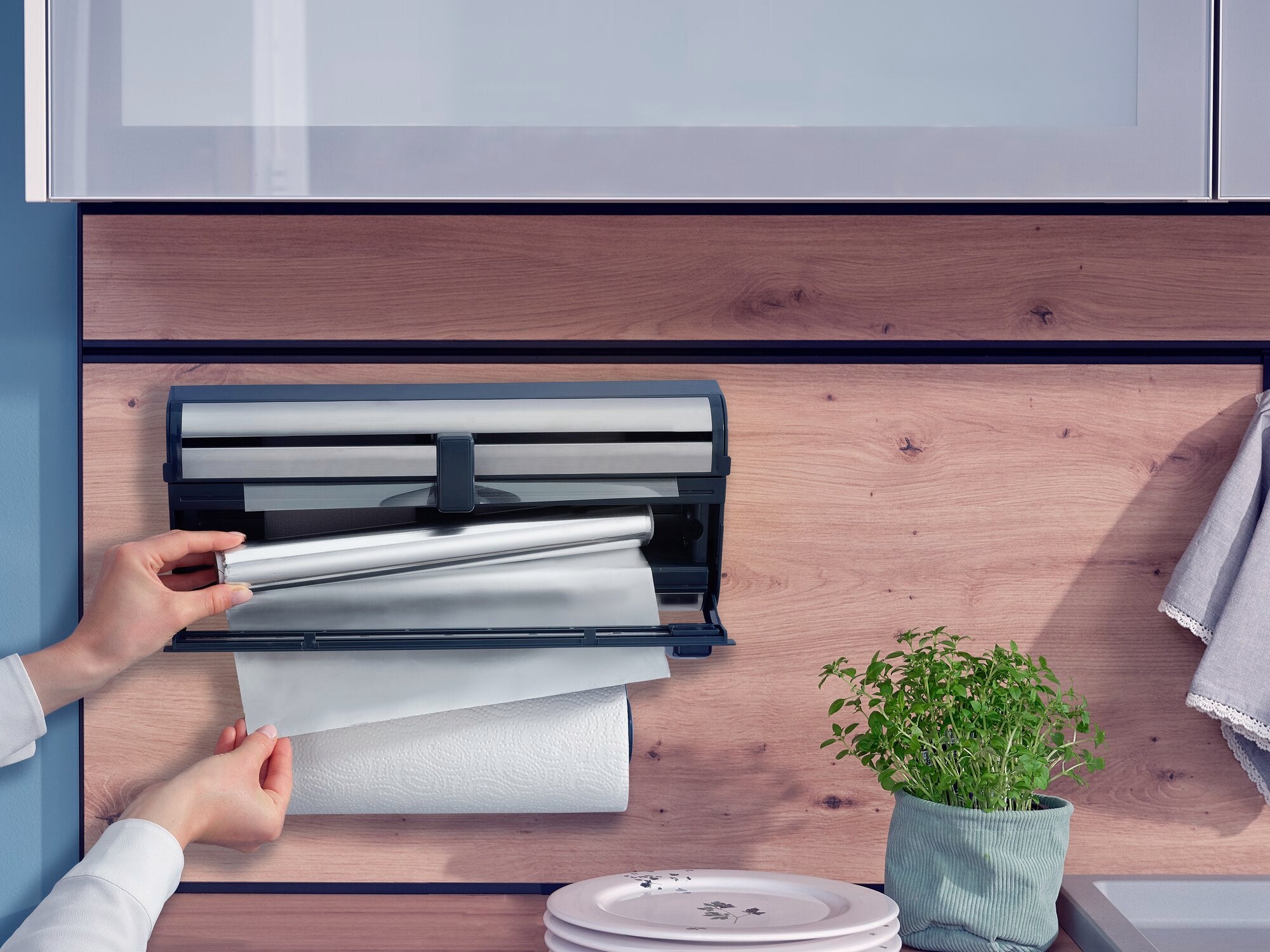 A Parat Royal wall-mounted roll holder is mounted on a wooden wall. A person pulls cling film from the holder. Kitchen rolls are visible underneath. Next to it is a pot of herbs.