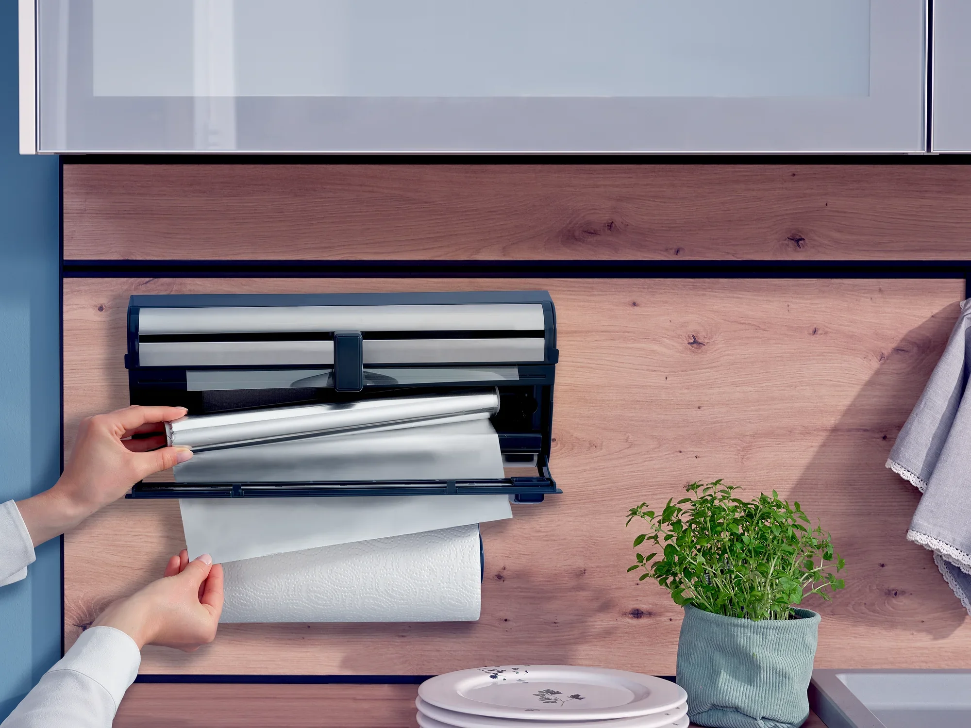 Uchwyt ścienny na rolki Parat Royal jest zamontowany na drewnianej ścianie. Osoba wyciąga folię spożywczą z uchwytu. Rolki kuchenne są widoczne pod spodem. Obok stoi doniczka z ziołami.