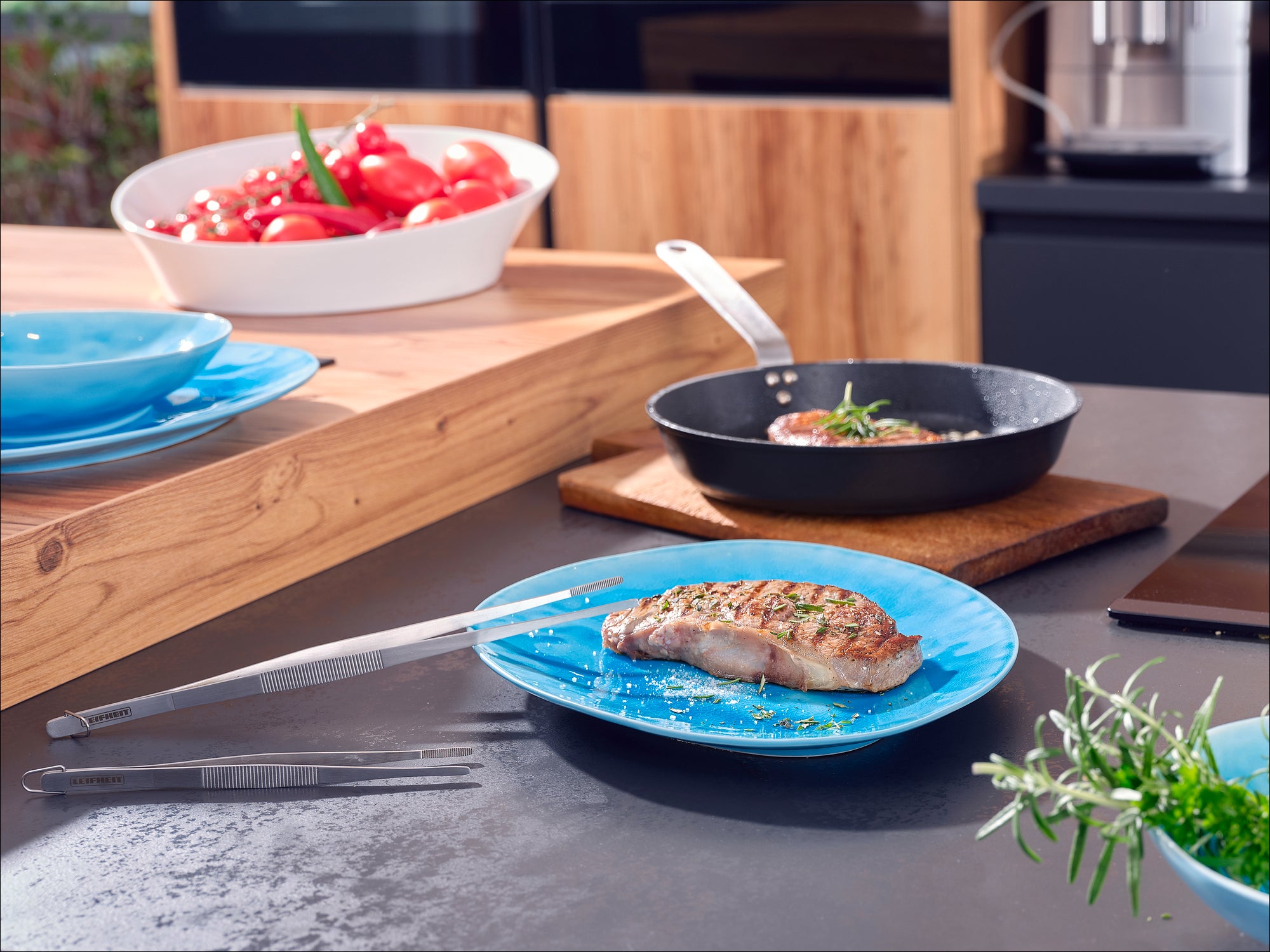 Ein modernes Küchenambiente mit einem blauen Teller, auf dem ein saftiges Steak liegt. Im Hintergrund eine Pfanne mit Kräutern und eine Schüssel mit frischen Tomaten.