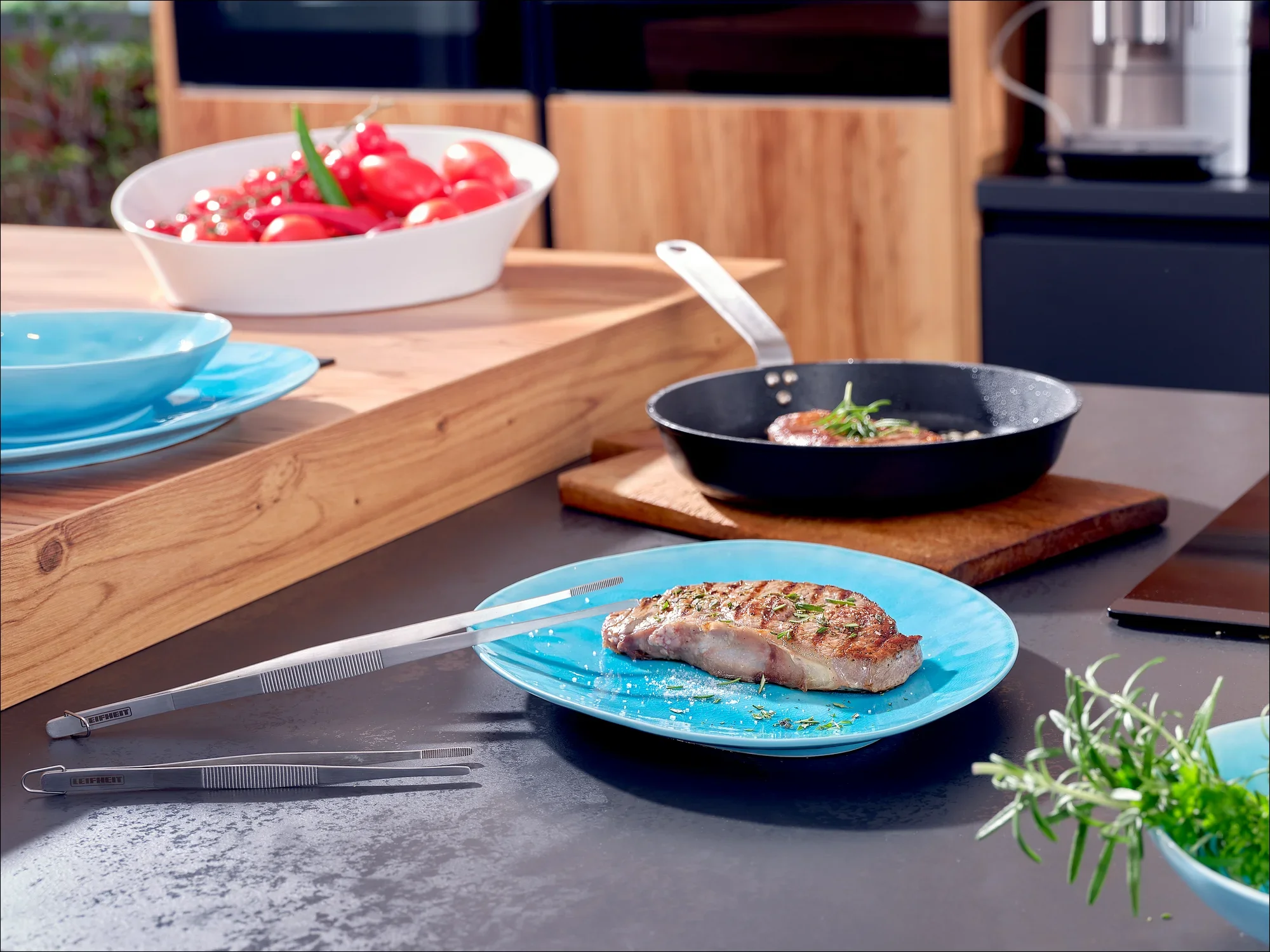 Nowoczesna atmosfera w kuchni z niebieskim talerzem trzymającym soczysty stek. W tle patelnia z ziołami i miska świeżych pomidorów.