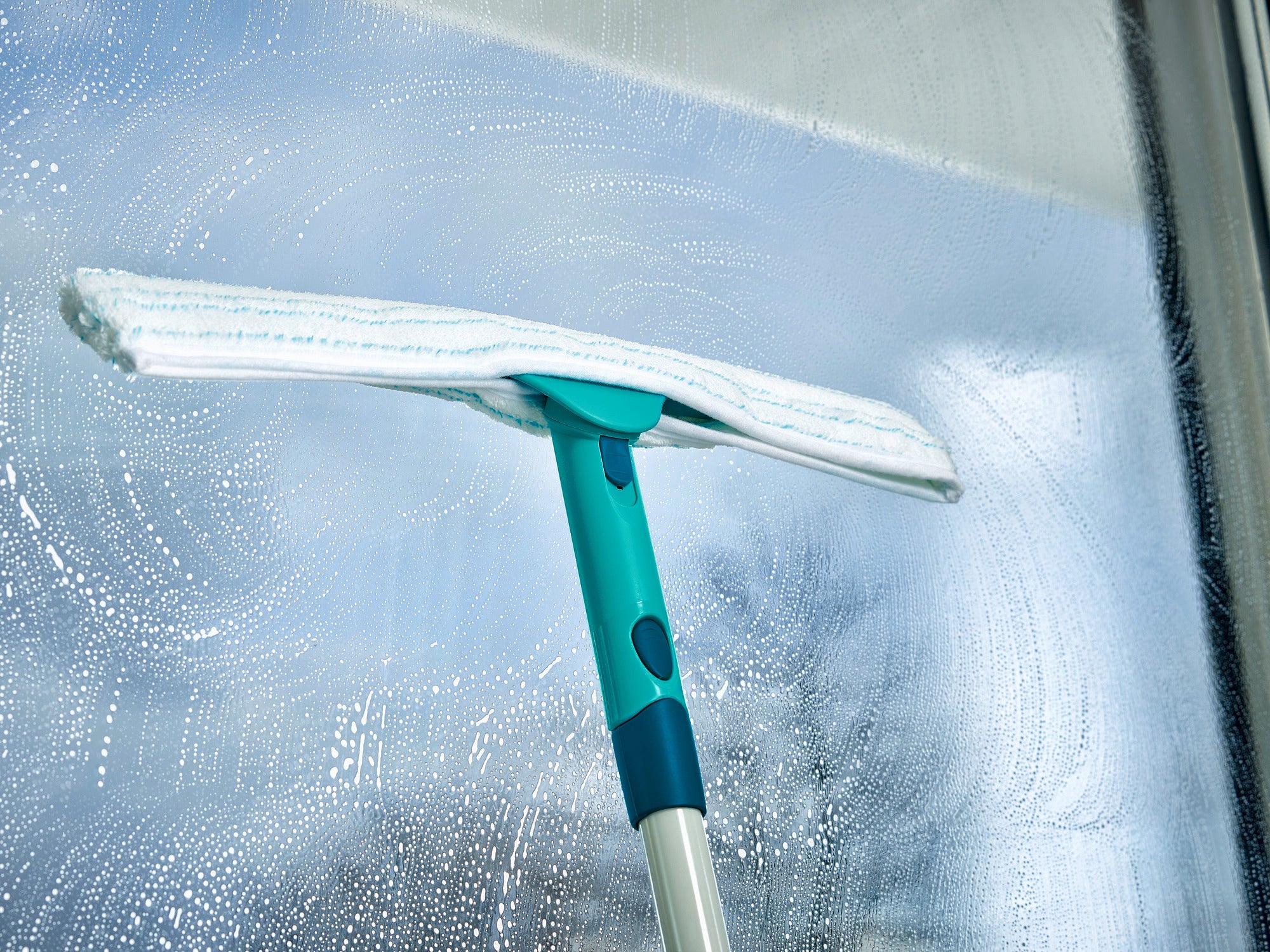 Fensterabzieher Window Slider M in Aktion: Ein türkisfarbener Fensterabzieher reinigt eine große Glasfläche mit kreisenden Bewegungen, während das Tageslicht durch das Fenster scheint.