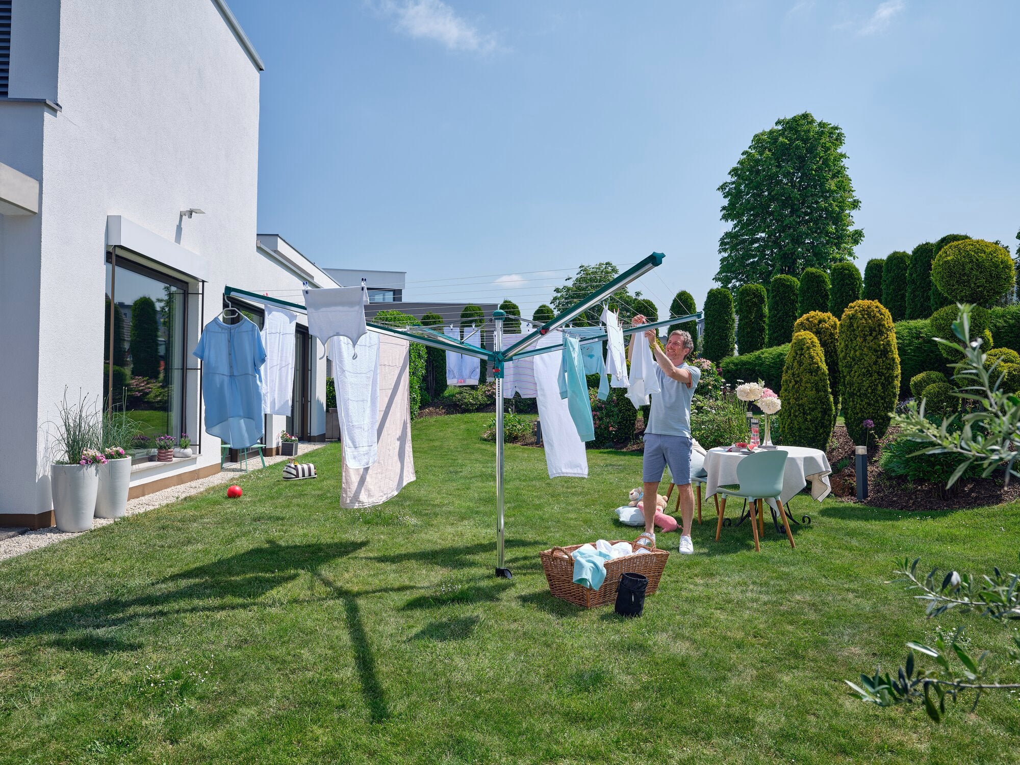 Ein Mann hängt Wäsche auf einem Linomatic 600 Deluxe Wäscheschirm im Garten auf. Der sonnige Tag und die grüne Umgebung schaffen eine angenehme Atmosphäre.