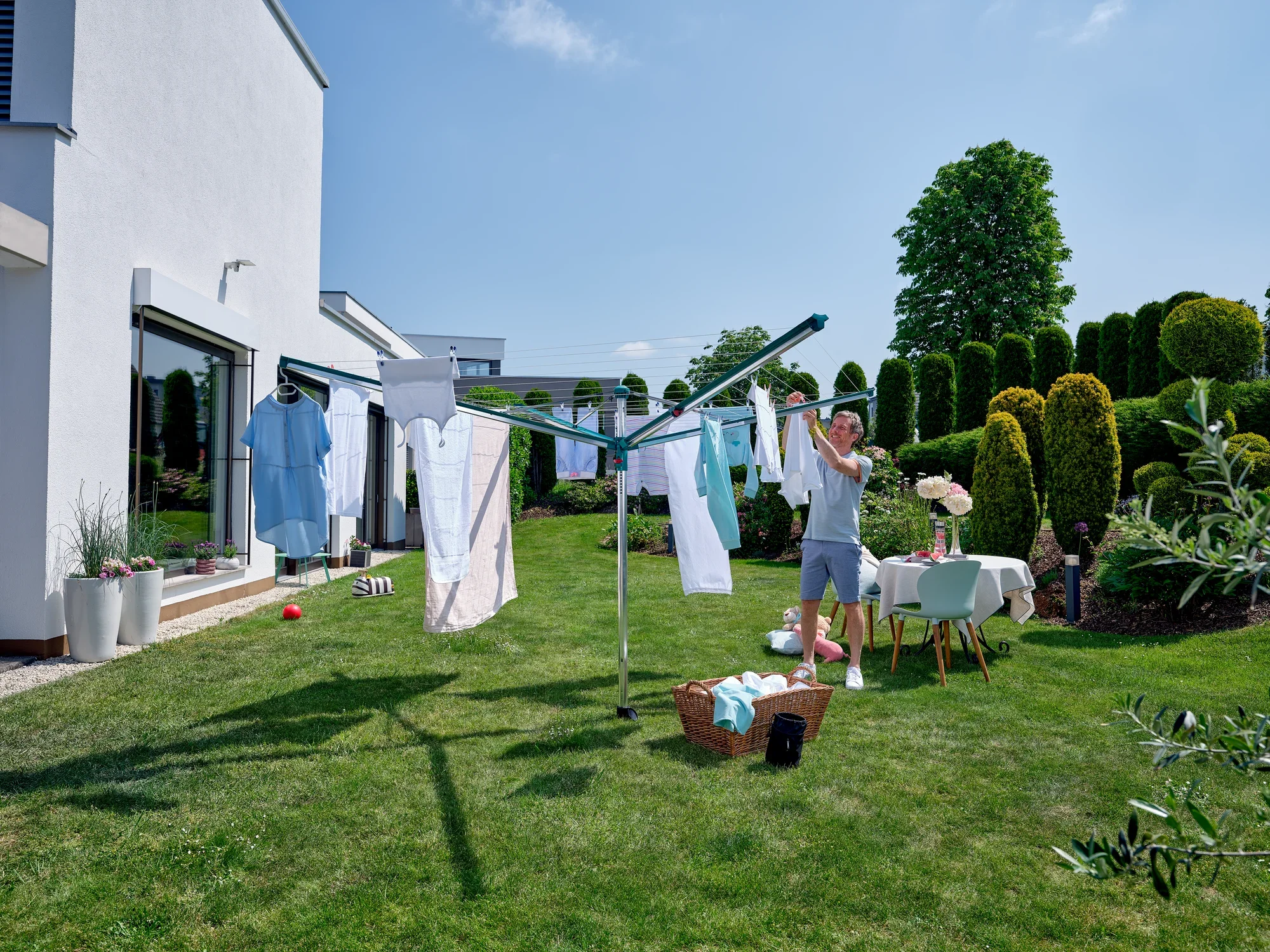 Mężczyzna wiesza pranie na parasolu Linomatic 600 Deluxe w ogrodzie. Słoneczny dzień i zielone otoczenie tworzą przyjemną atmosferę.