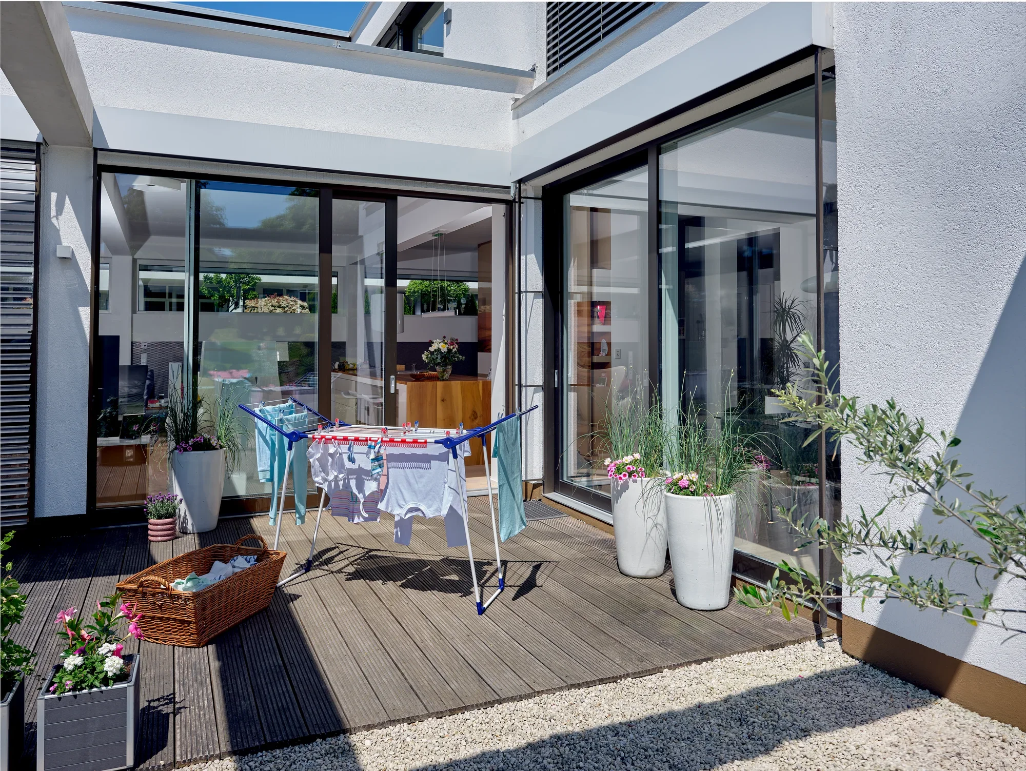 Ein moderner Standtrockner Pegasus 150 Solid Slim steht auf einer sonnigen Terrasse. Er ist mit bunter Wäsche behängt. Umgeben von Pflanzen und einem Korb mit Wäscheklammern.