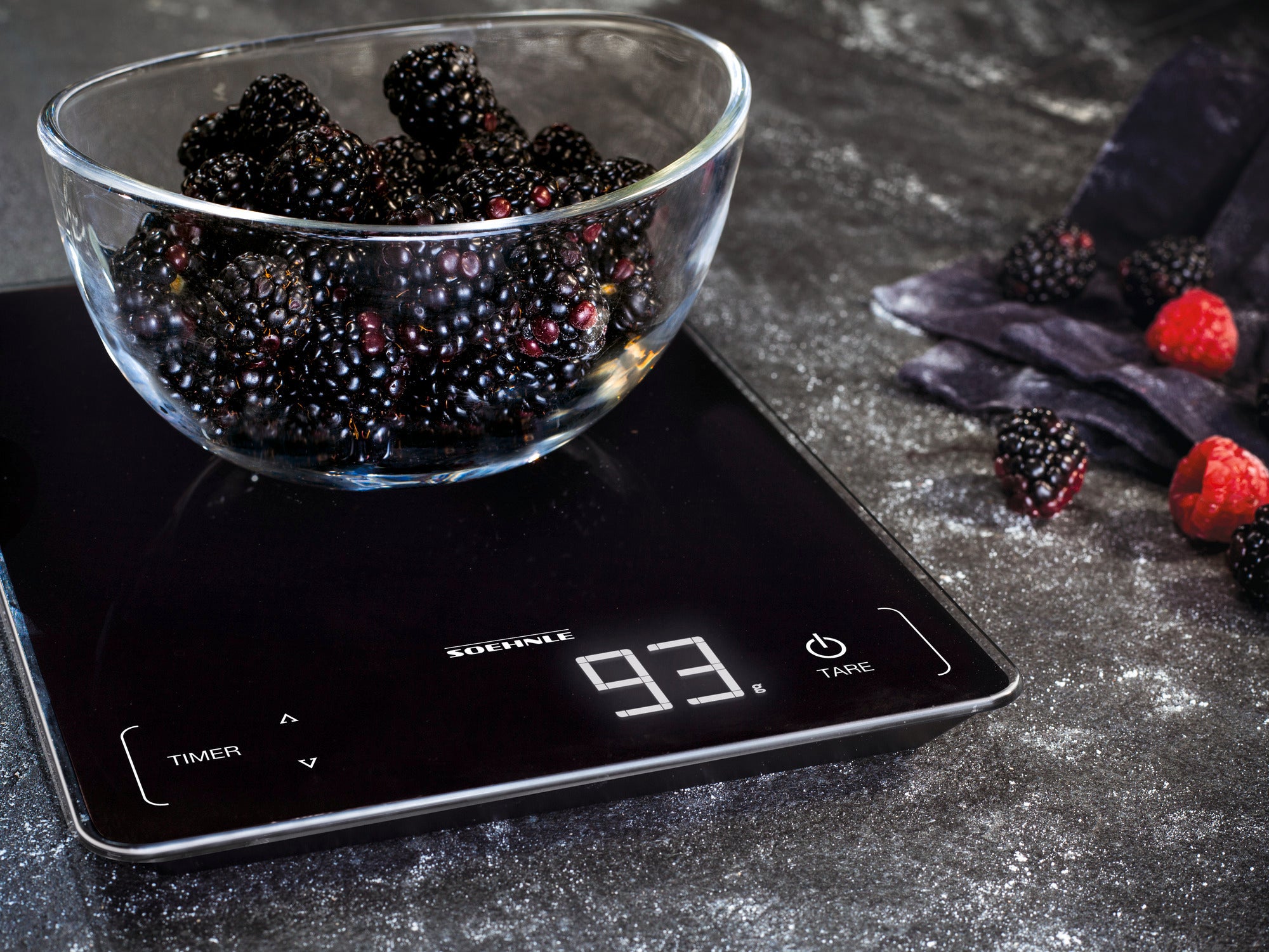 Eine digitale Küchenwaage zeigt das Gewicht einer Schale voller Brombeeren an. Die Waage hat ein modernes Design mit einem klaren, digitalen Display, das 93 Gramm anzeigt.