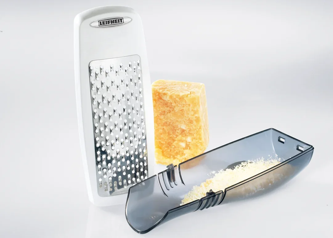 Une râpe à parmesan Leifheit à côté d'un morceau de parmesan et d'un bol de fromage râpé. La râpe a un design moderne et est idéale pour la cuisine.
