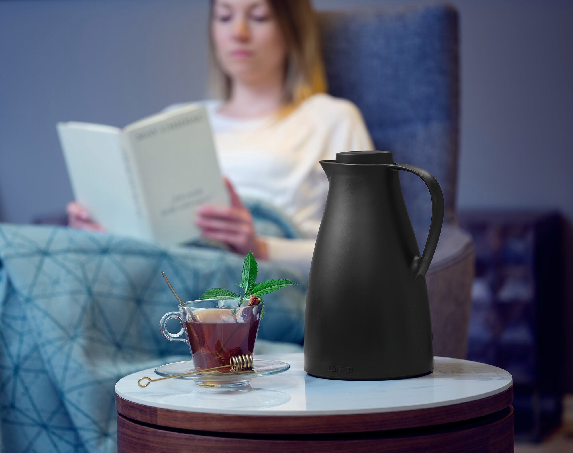 Eine schwarze Isolierkanne namens Harmonic steht auf einem kleinen Tisch. Daneben eine Tasse Tee mit Minzblättern. Im Hintergrund liest eine Person ein Buch auf einem Sessel.