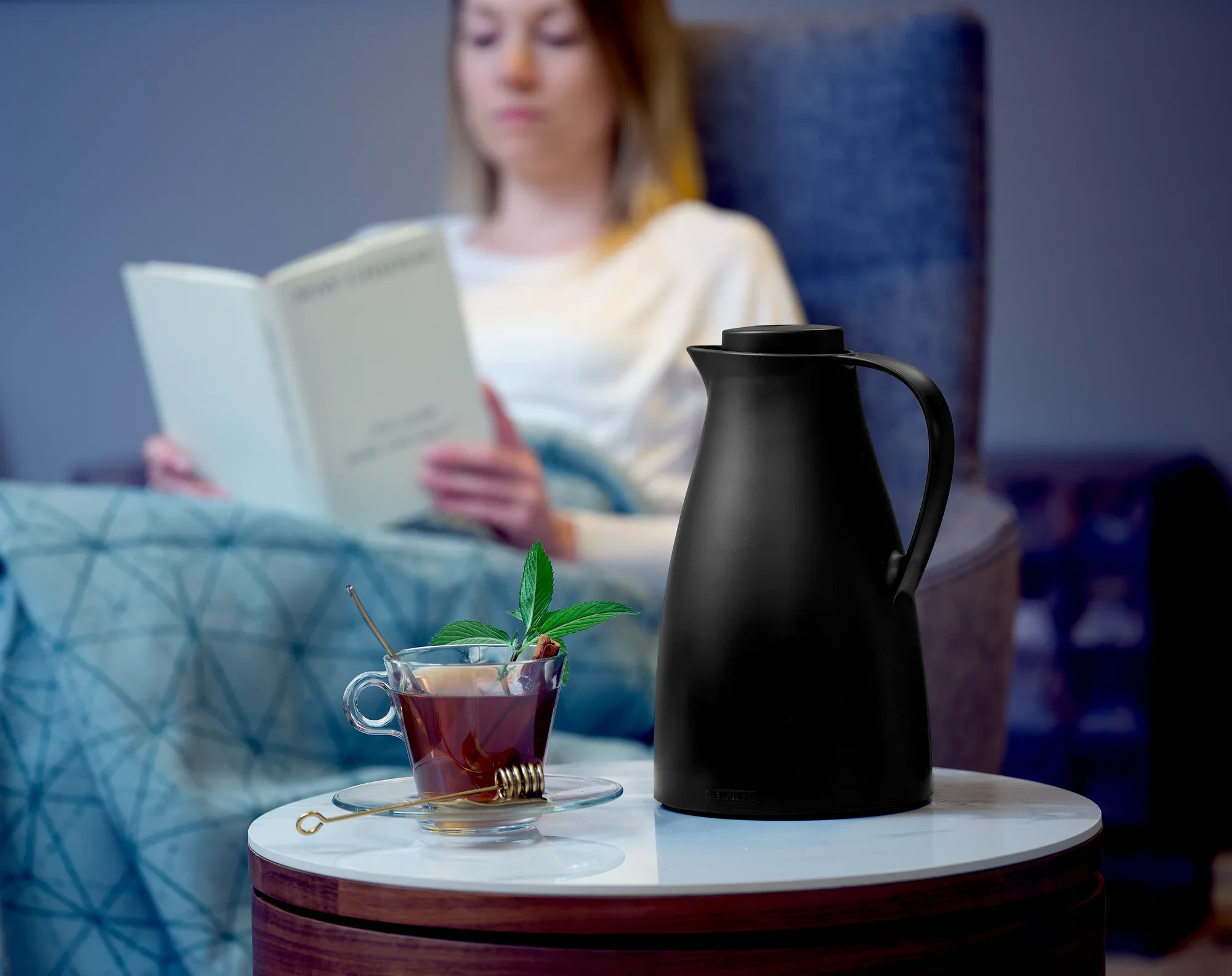 Una jarra térmica negra llamada Harmonic está sobre una mesita. Junto a ella, una taza de té con hojas de menta. Al fondo, una persona lee un libro en un sillón.