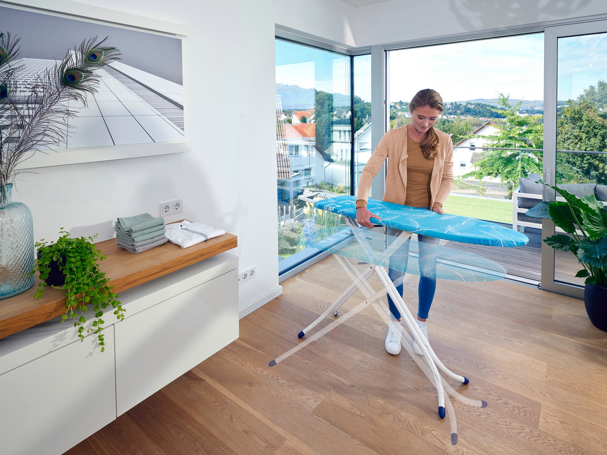 Eine Frau steht in einem modernen, hellen Raum und benutzt den Bügeltisch Air Board M Compact. Der Tisch ist leicht und hat ein modernes Design, ideal für effizientes Bügeln.