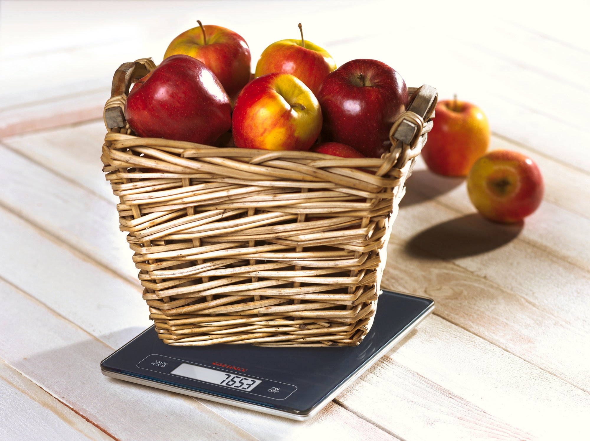 Een mand vol rode en gele appels staat op een digitale keukenweegschaal. De weegschaal geeft het gewicht van de appels aan en rust op een lichtgekleurd houten oppervlak.