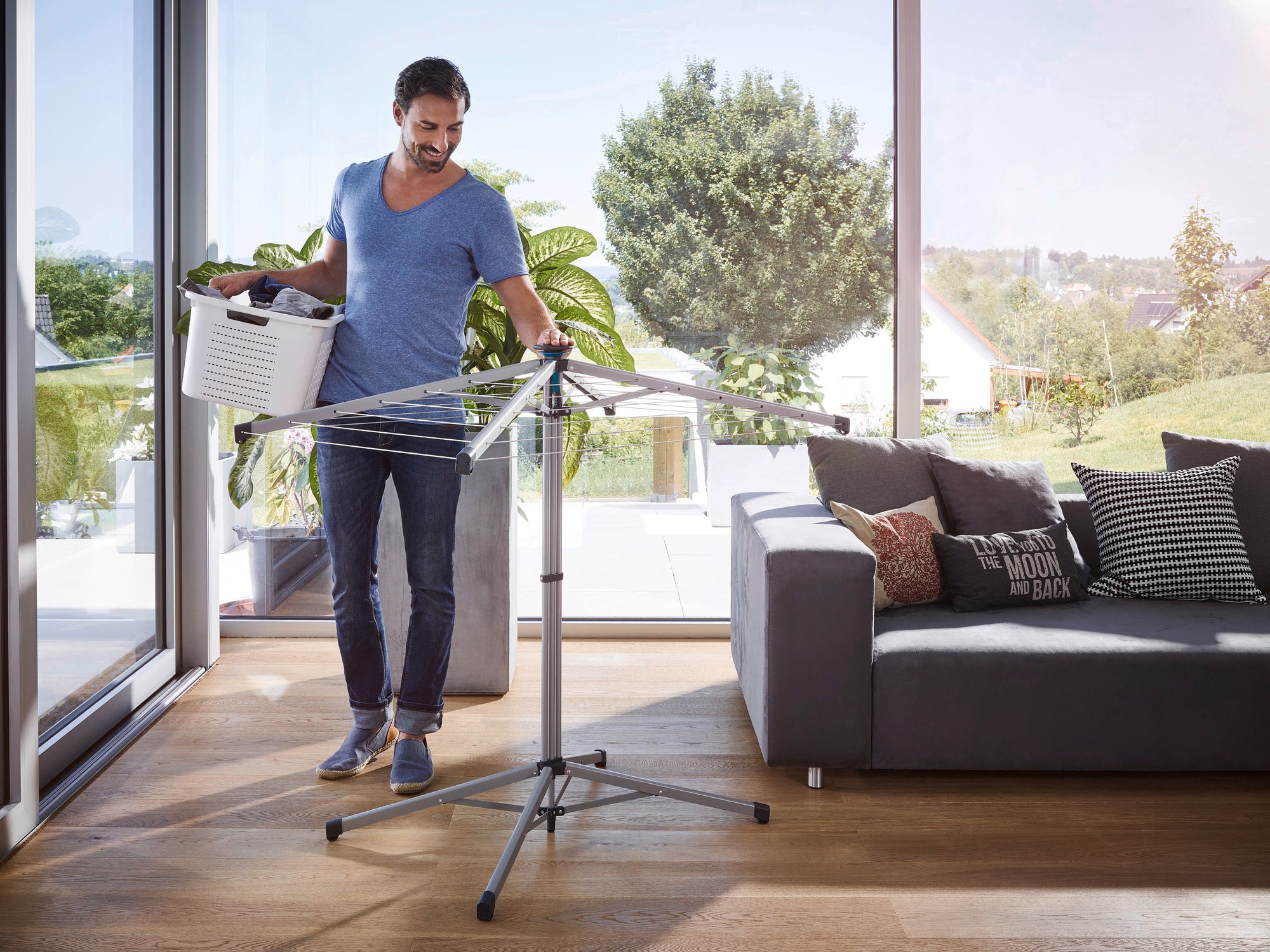 Ein Mann steht in einem modernen Wohnzimmer und benutzt die Standwäschespinne Lino Pop-Up. Er hält einen Wäschekorb und die Spinne ist kompakt und platzsparend.