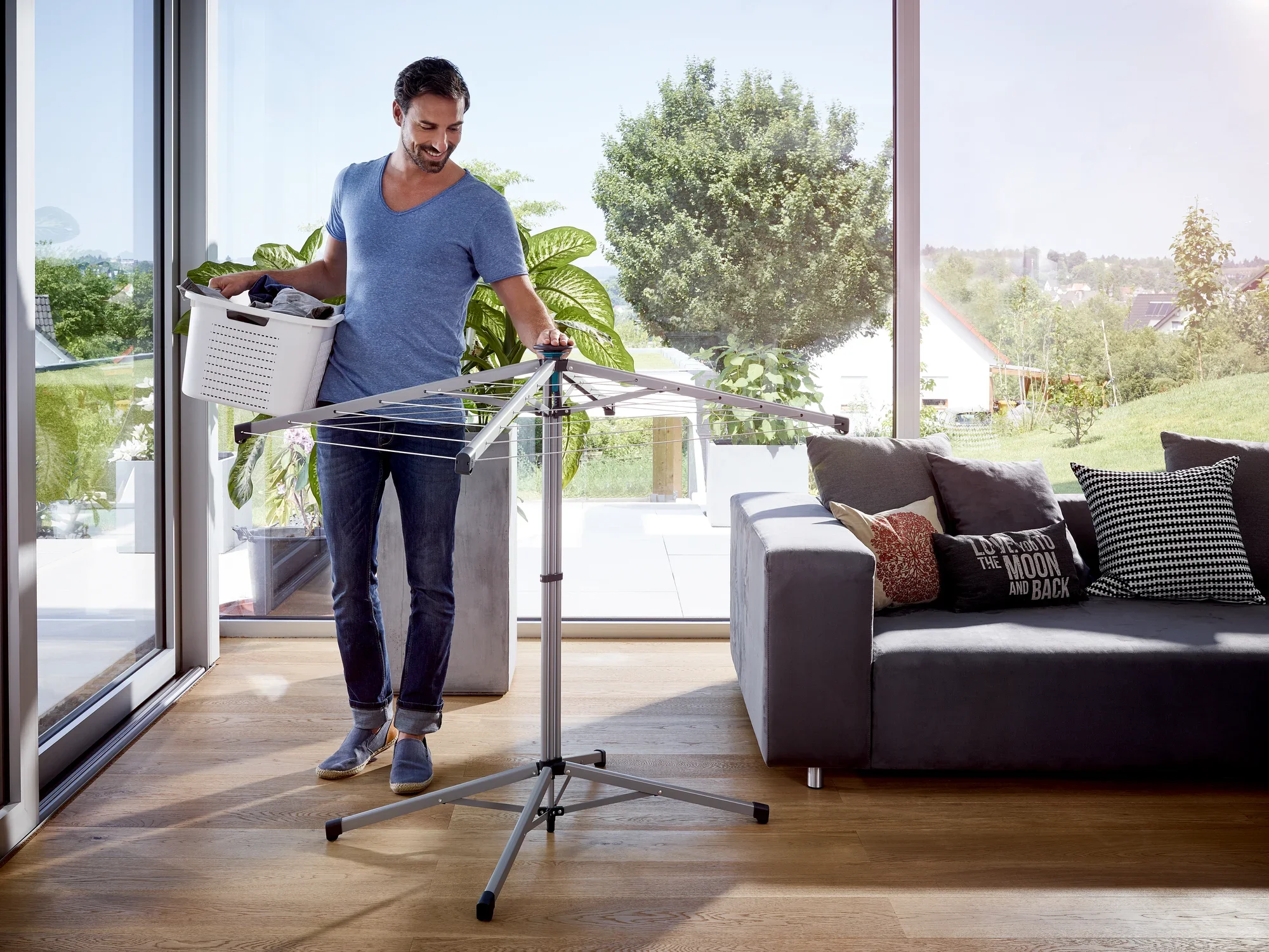 Un hombre está de pie en una moderna sala de estar y utiliza el tendedero de pie Lino Pop-Up. Sostiene una cesta de la ropa y el tendedero es compacto y ocupa poco espacio.