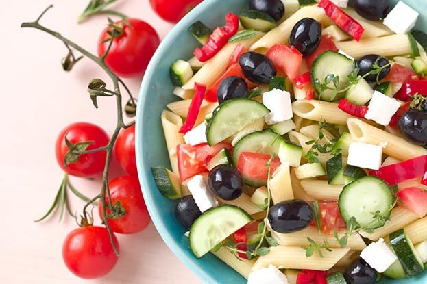 Een kleurrijke pastasalade met penne, tomaten, komkommers, olijven, paprika en feta in een blauwe schaal, met daarnaast verse tomaten aan de tros.