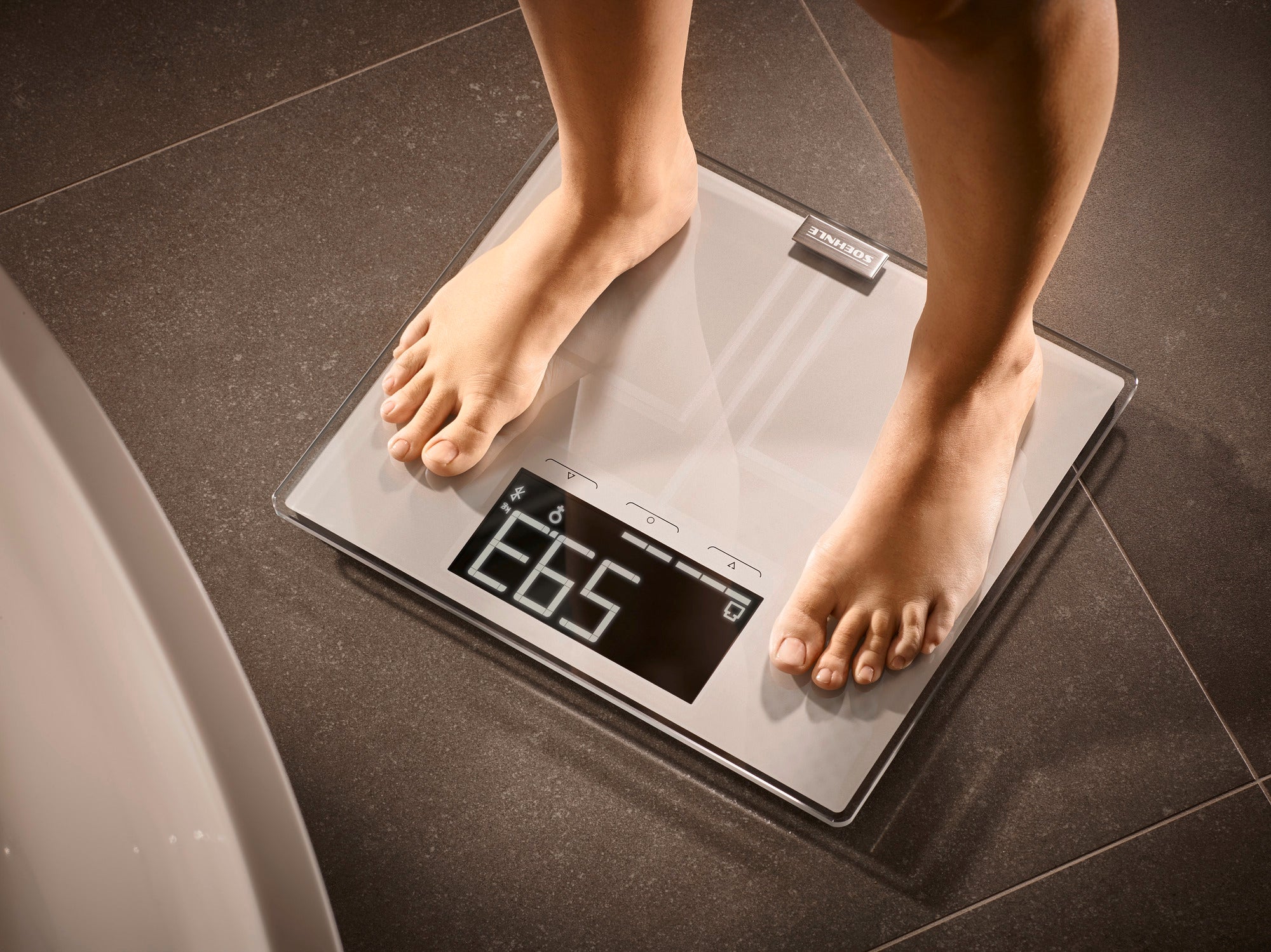 A person stands barefoot on the Shape Sense Connect 200 digital bathroom scale. The scale displays the weight and has a modern, transparent design.