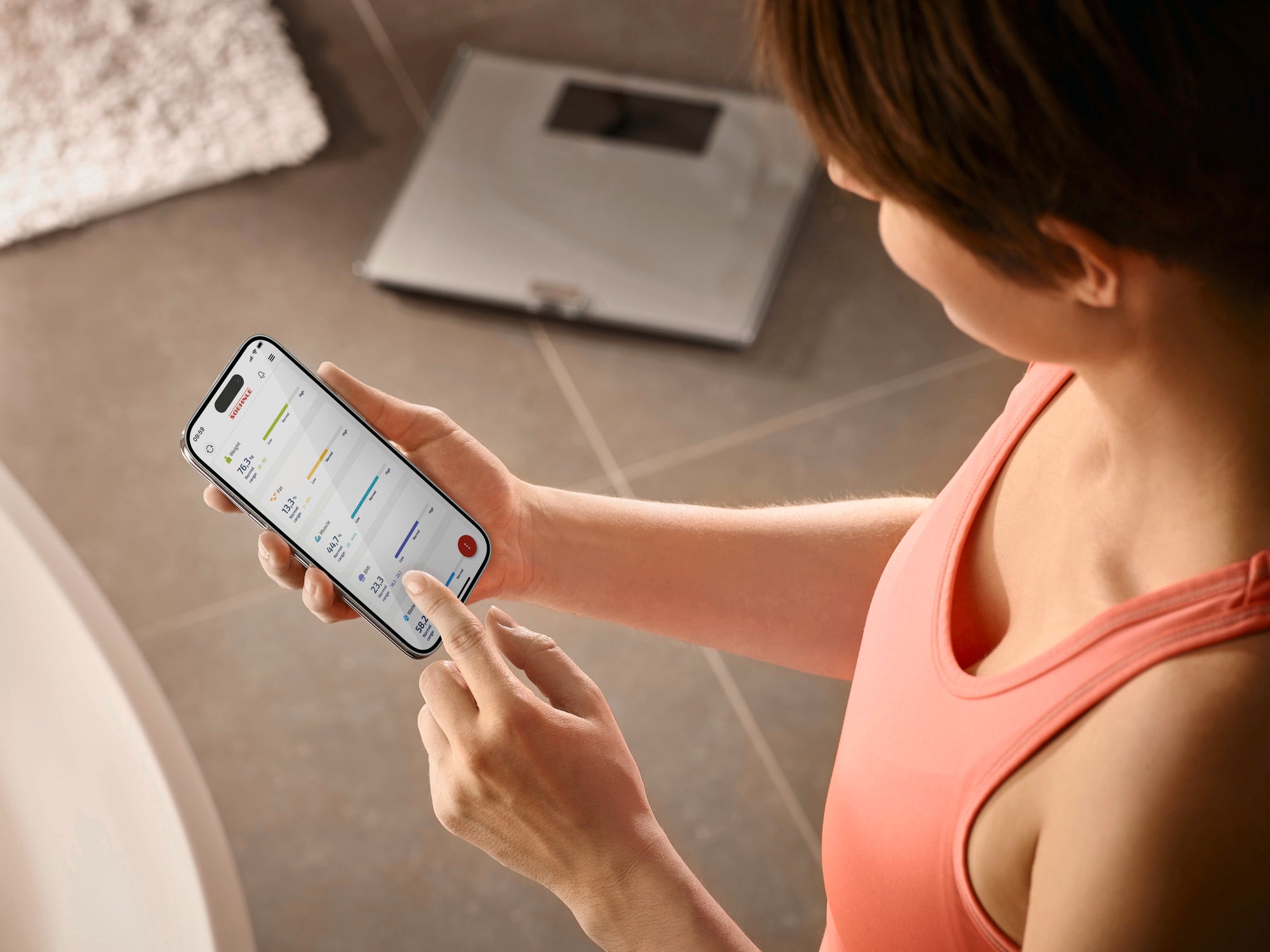 Eine Person verwendet die Personenwaage digital Shape Sense Connect 200. Sie überprüft die Messwerte auf ihrem Smartphone, während die Waage im Hintergrund sichtbar ist.