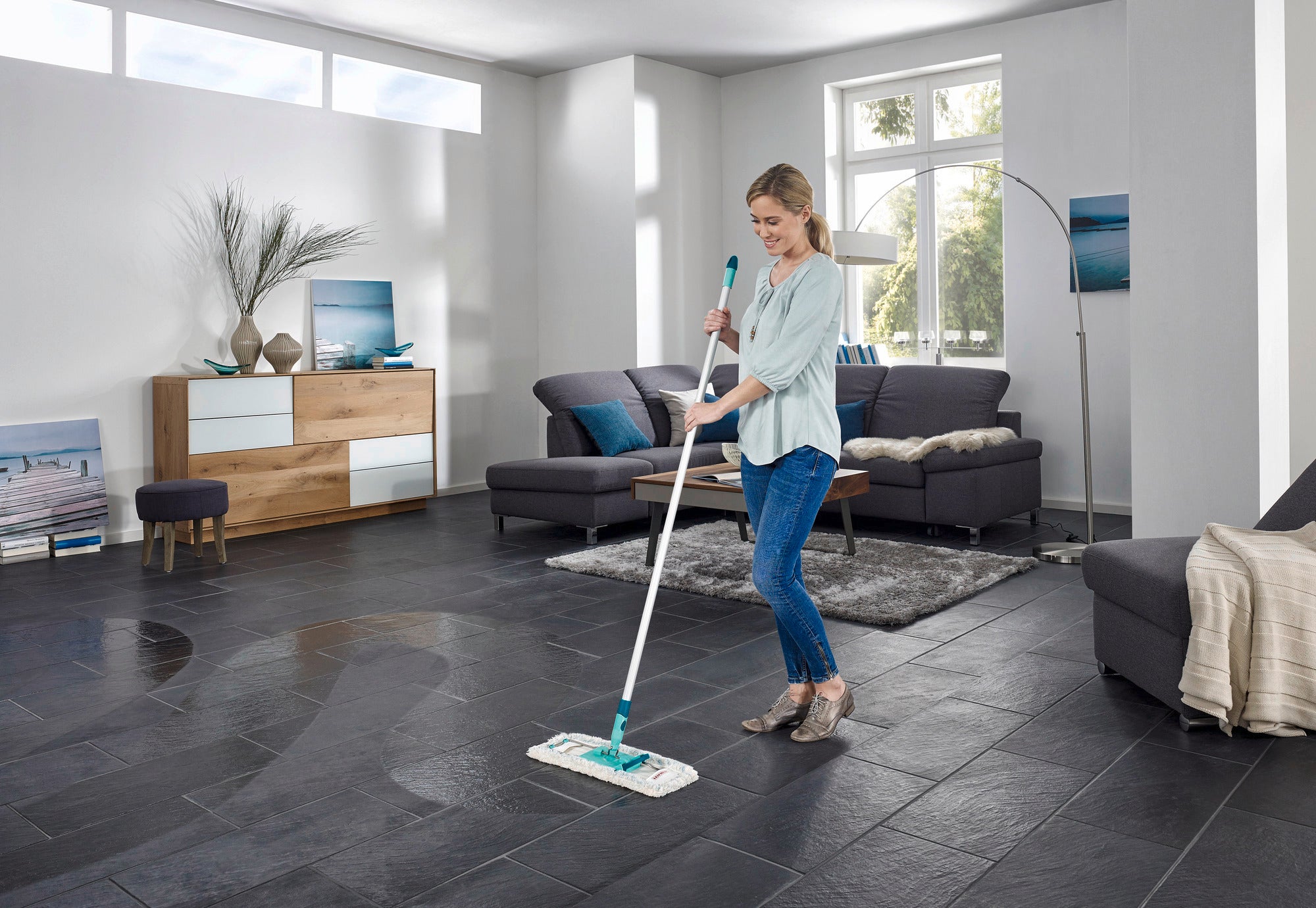 Une femme nettoie une grande pièce moderne avec un sol en carrelage foncé à l'aide du balai Profi XL. La pièce est bien éclairée et meublée avec goût, avec un canapé et des décorations.