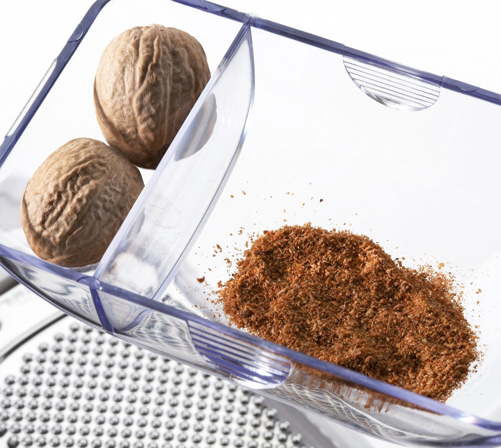 A transparent container holds two whole nutmegs and a pile of grated nutmeg, with a grater partially visible underneath.