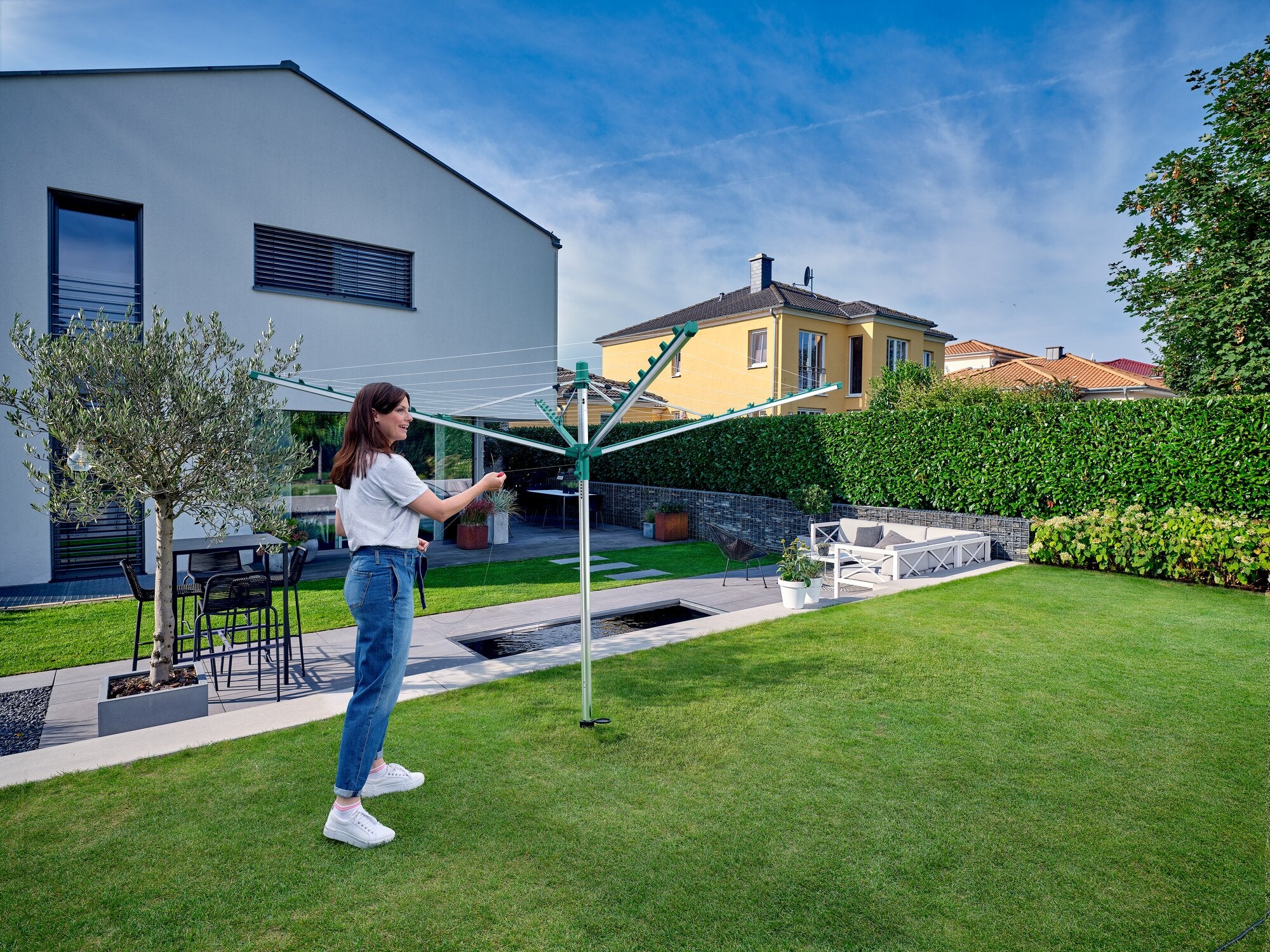 Eine Frau steht in einem gepflegten Garten und öffnet eine Wäschespinne Linomatic V 400. Im Hintergrund sind moderne Häuser und eine Hecke zu sehen. Der Himmel ist klar und blau.