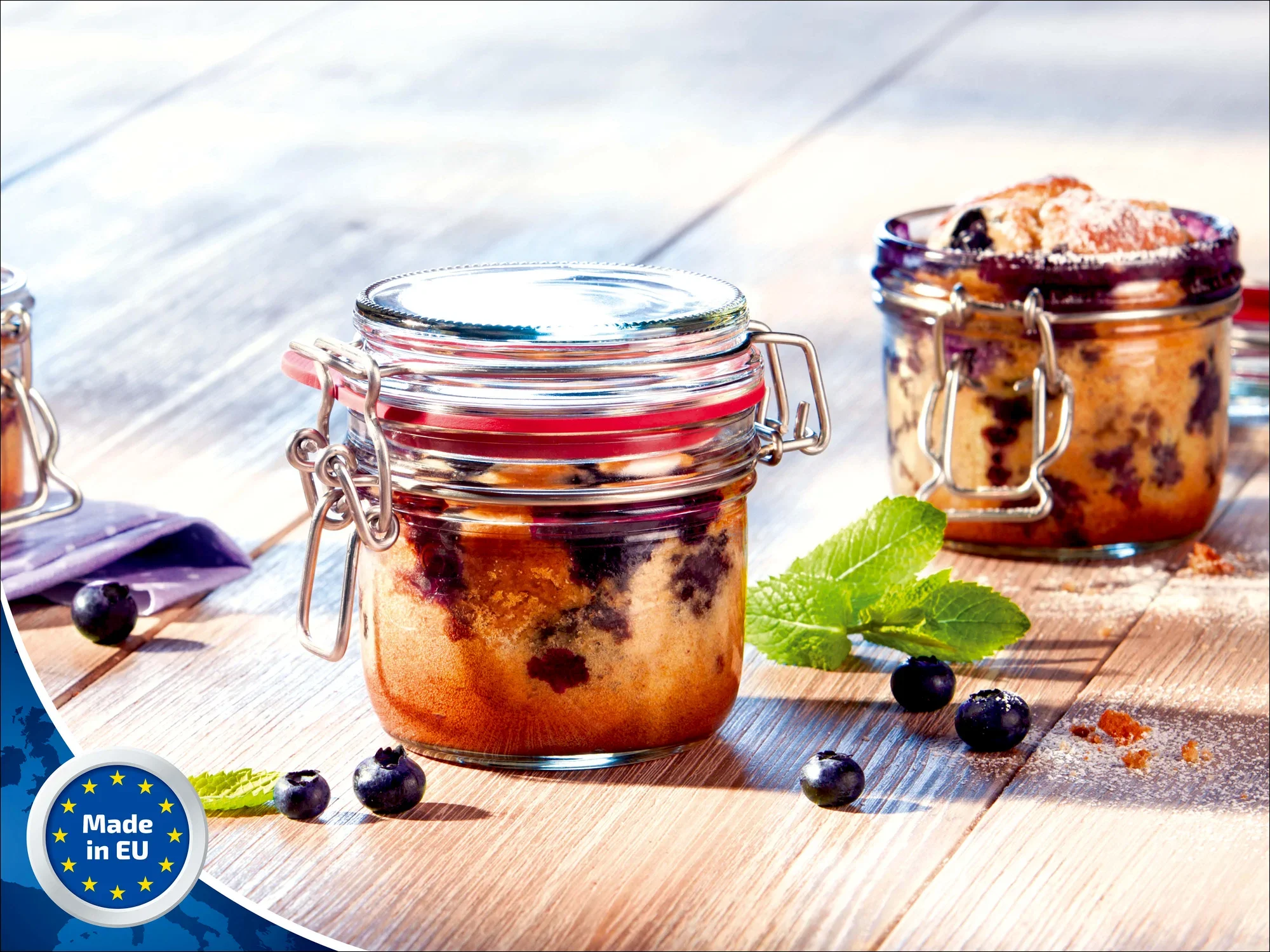 Ein Drahtbügelglas von Leifheit mit 135 ml Fassungsvermögen, gefüllt mit einem Dessert, steht auf einem Holztisch. Daneben liegen frische Blaubeeren und ein Minzblatt.