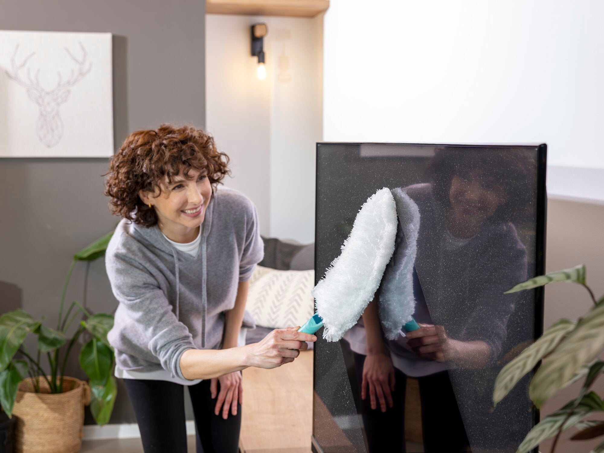 Eine Person verwendet den SUPERDUSTER XL Staubwedel, um einen großen Bildschirm zu reinigen. Der Staubwedel hat einen weißen, flauschigen Kopf und einen blauen Griff.